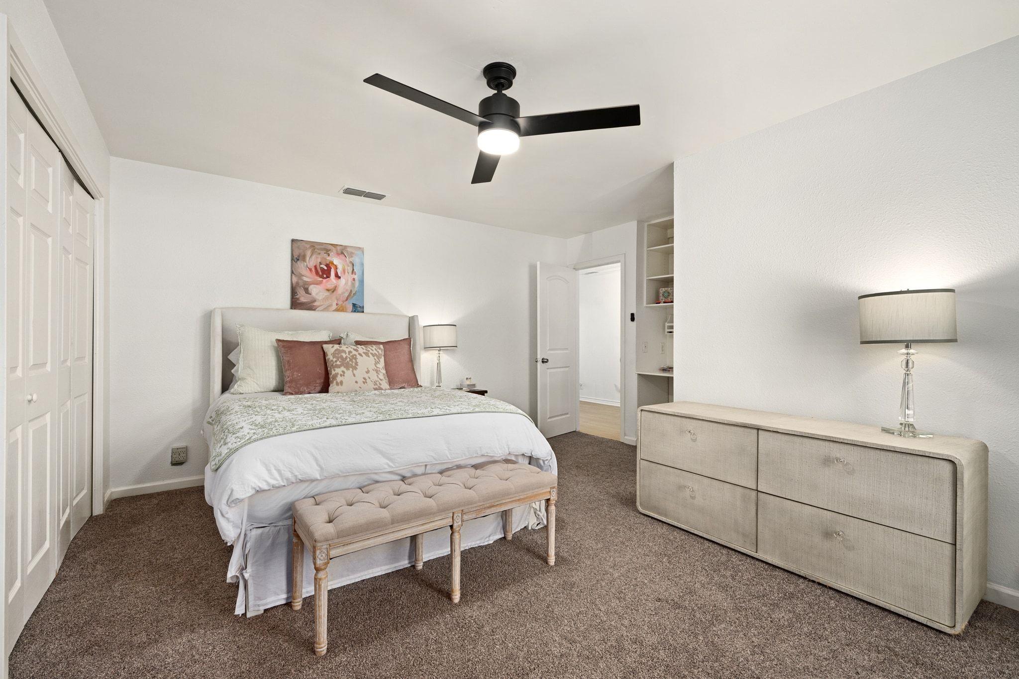 Staged bedroom with an upholstered bed, tufted bench, ceiling fan, and long dresser.
