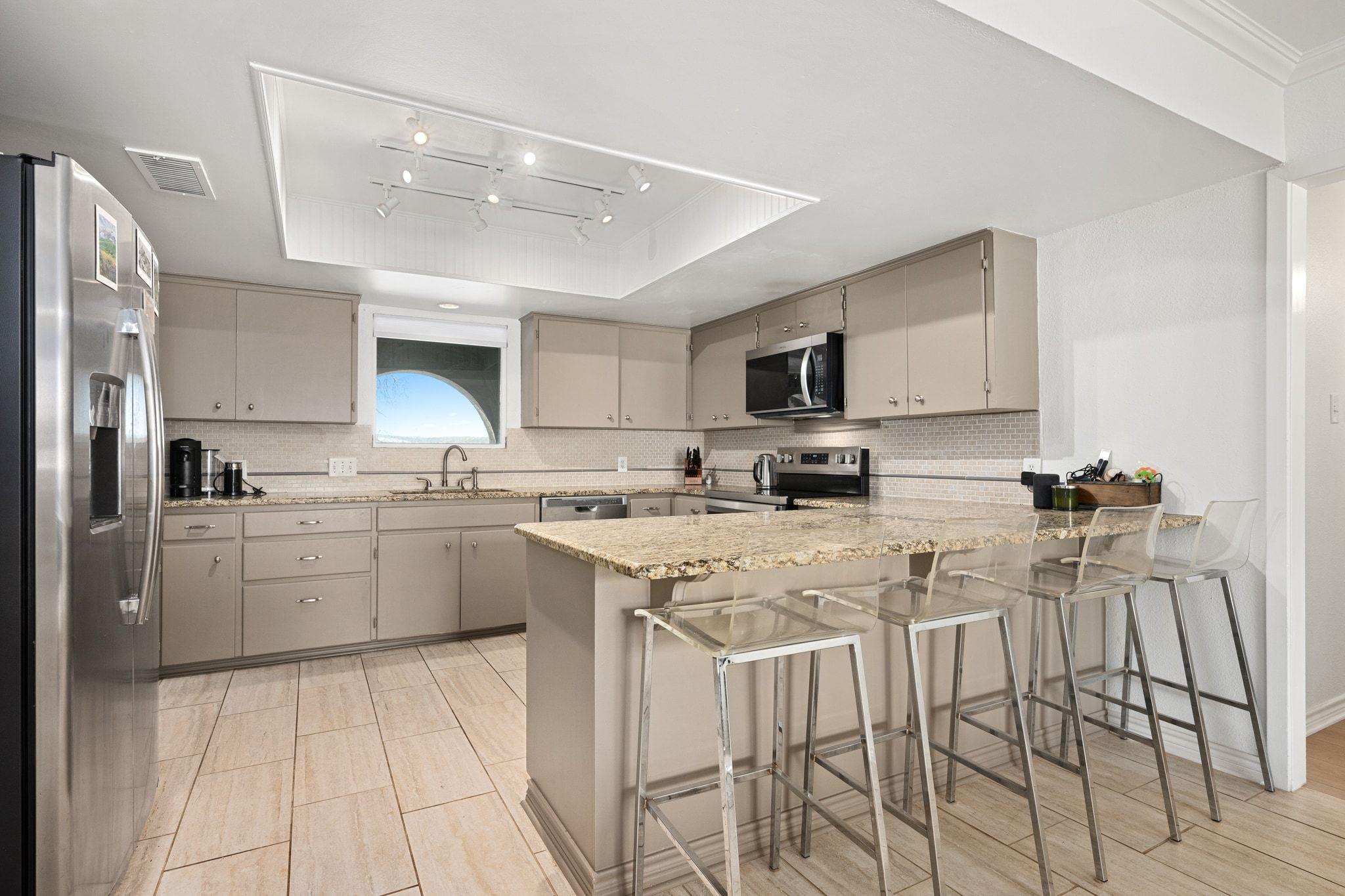 Kitchen with a granite island, clear bar stools, taupe cabinets, and stainless steel appliances.
