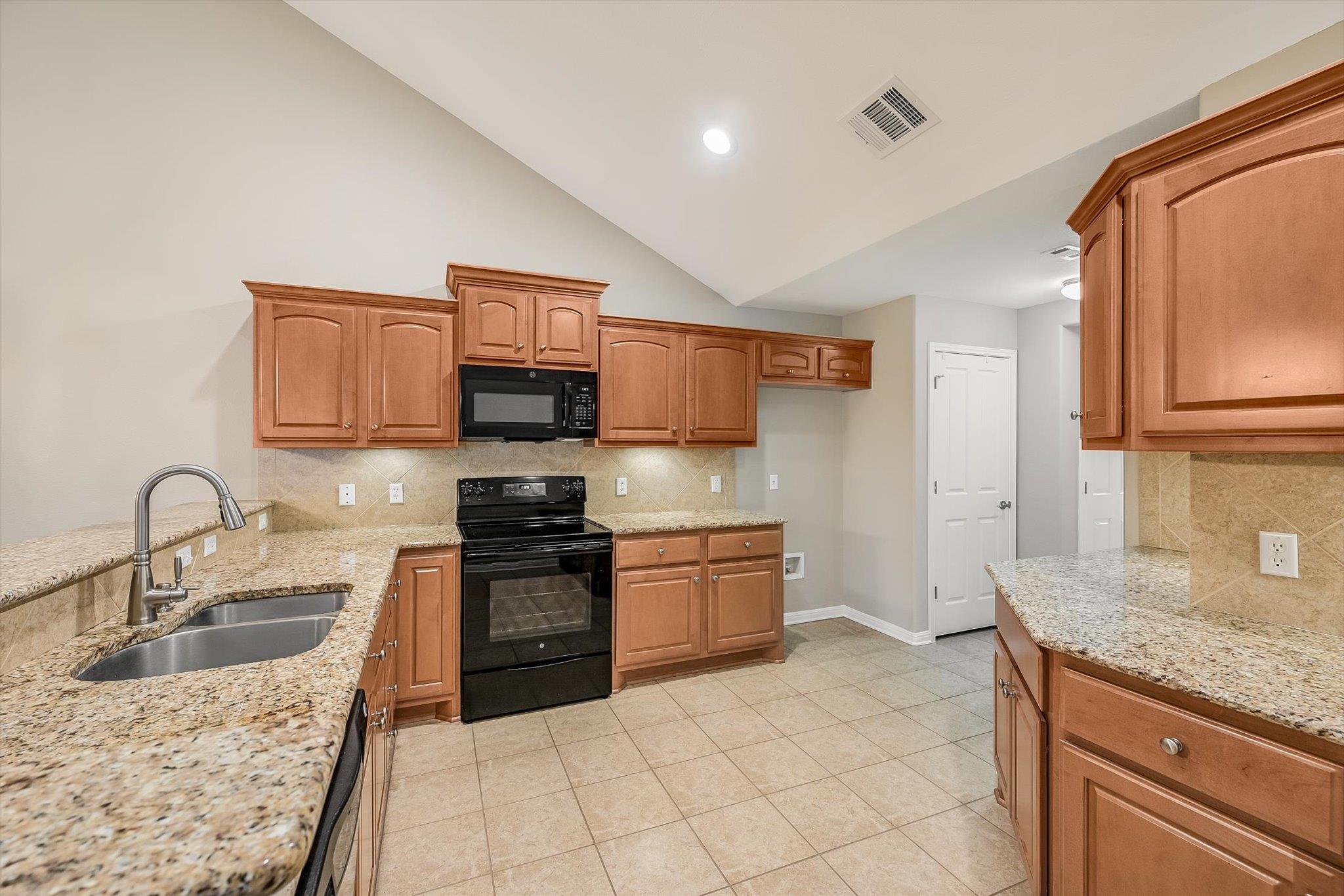 Kitchen with warm wood cabinets, granite countertops, and black appliances beneath a vaulted ceiling.