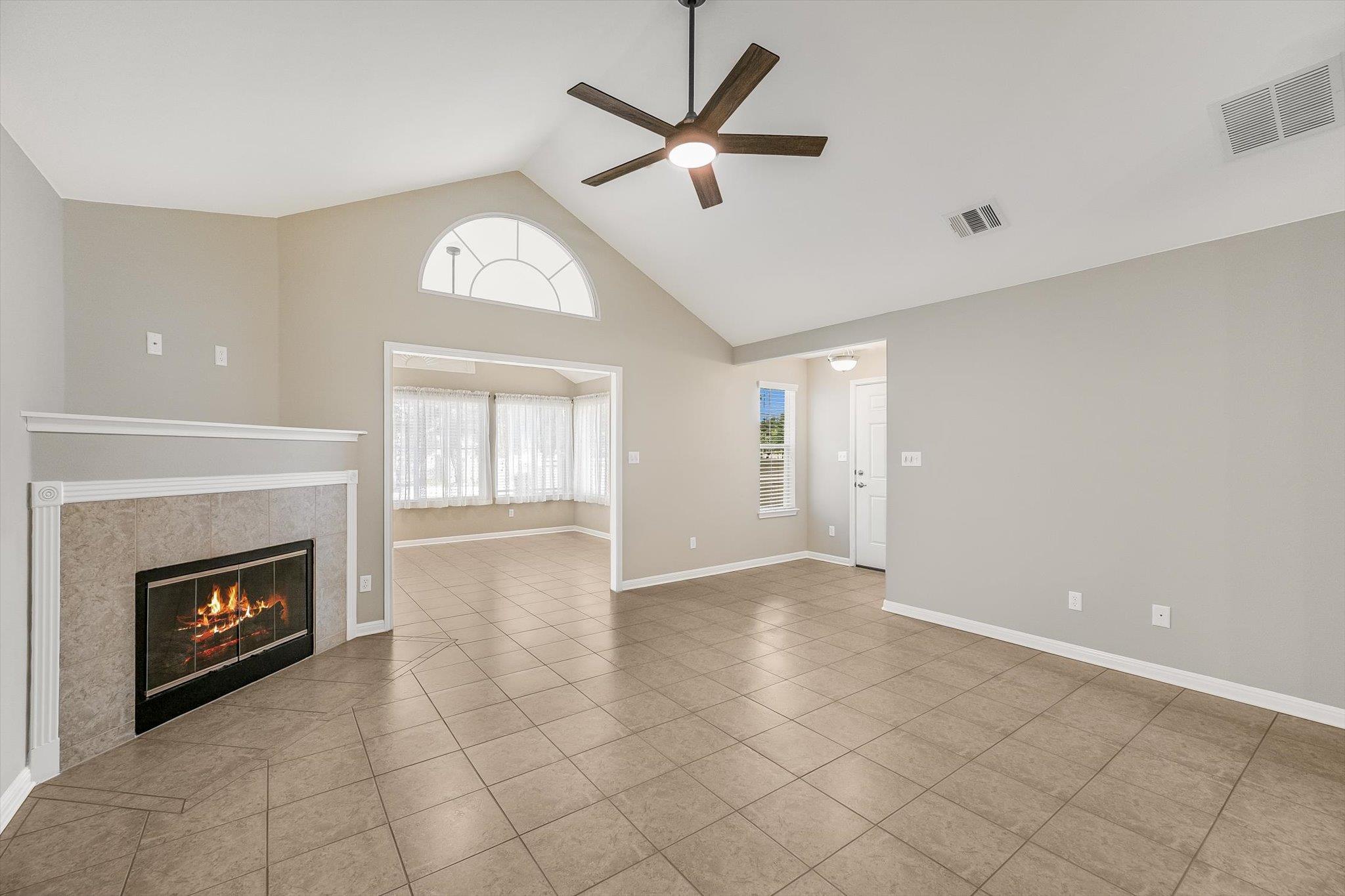 Empty living room with a vaulted ceiling, tiled fireplace, and wide opening to a bright adjoining room.