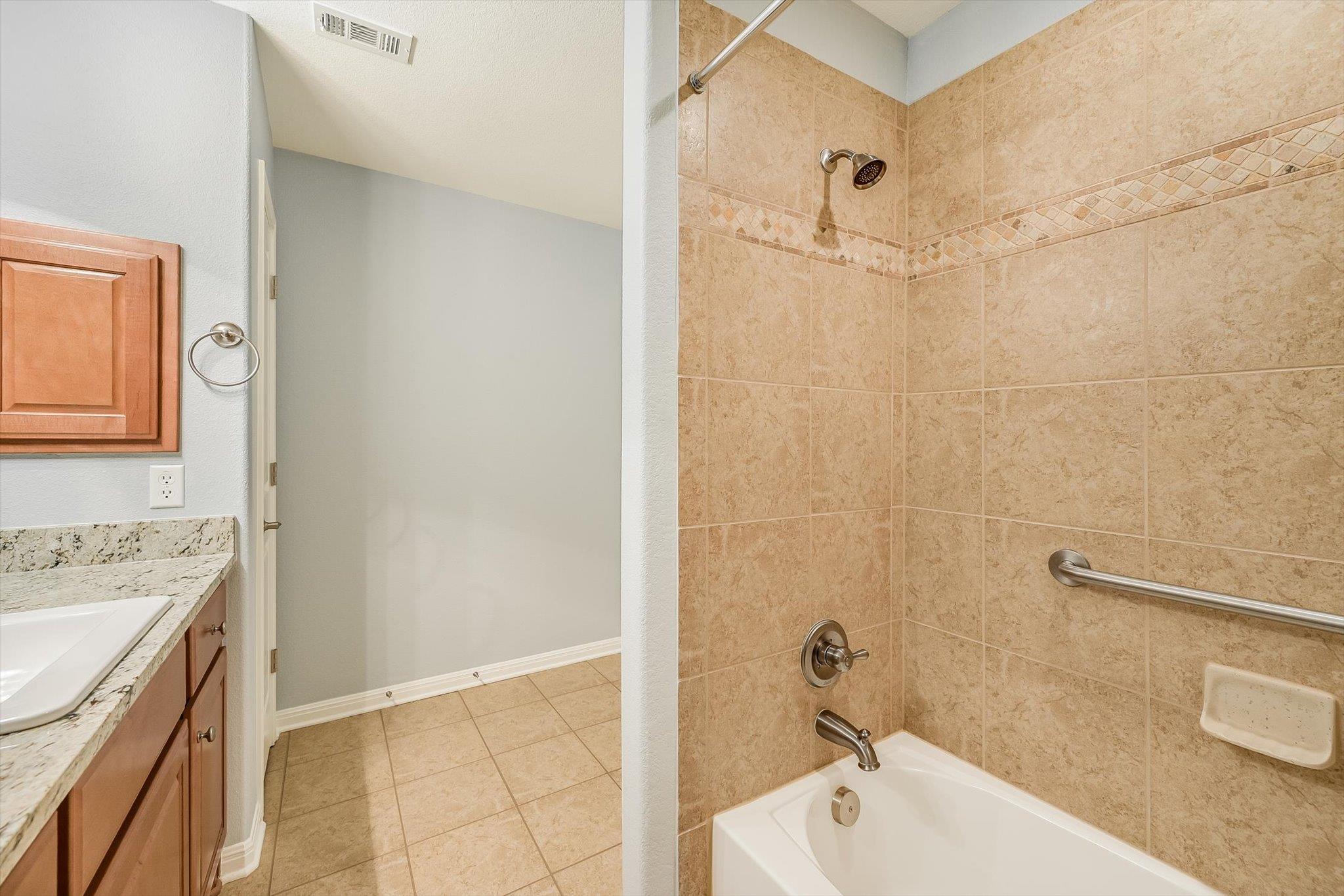 Bathroom with a tan tiled tub and shower, grab bar, and wood vanity with granite countertop.