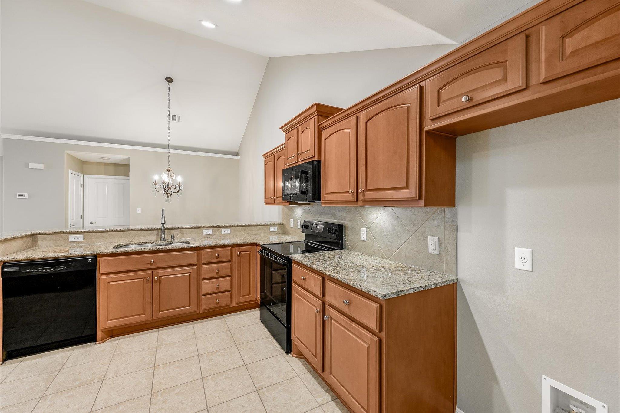 Kitchen with warm wood cabinets, granite counters, black appliances, and a chandelier above the sink.