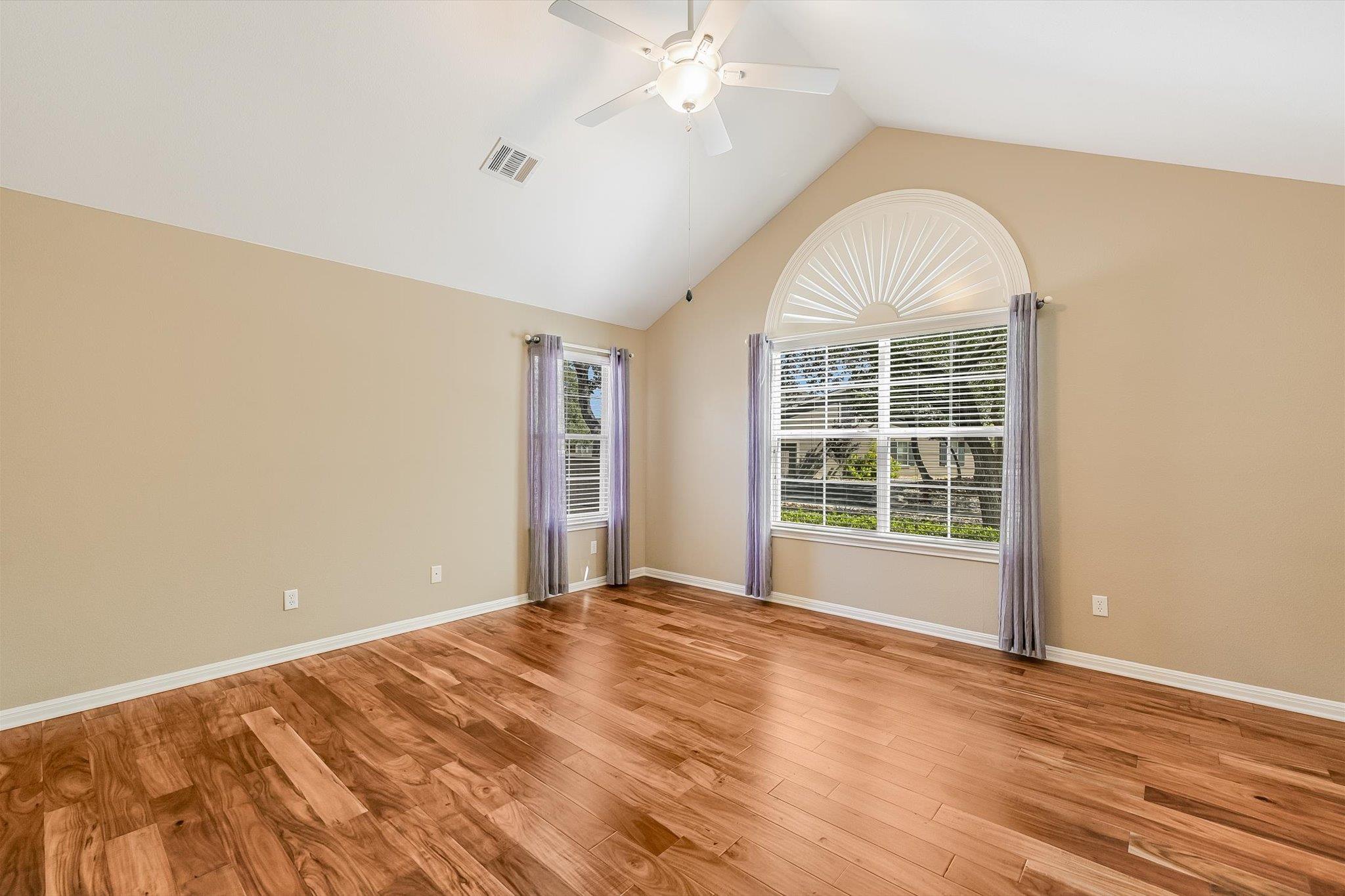 Vacant room with wood plank flooring, vaulted ceiling, and a large arched window with blinds.