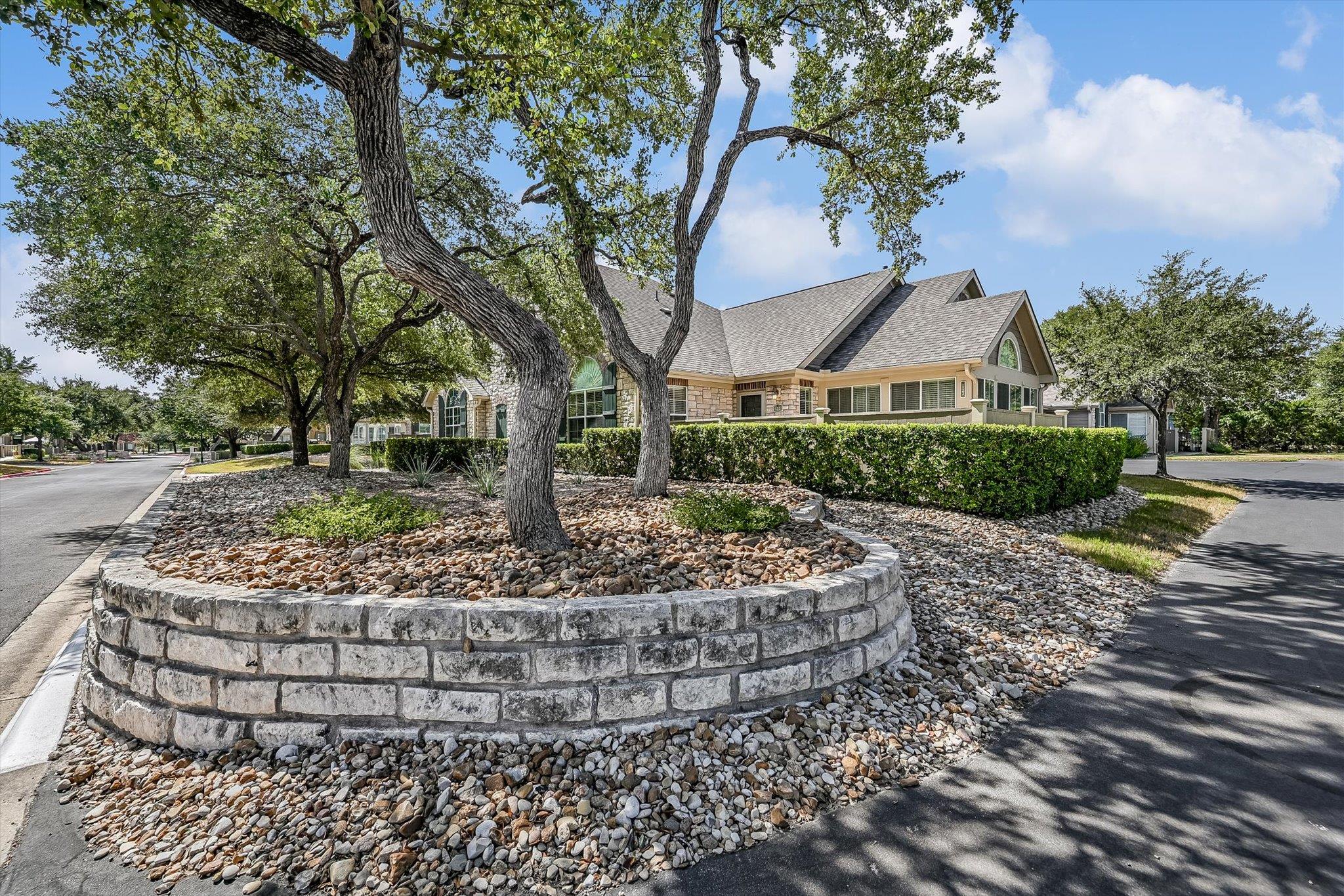 Stone-and-siding home behind mature oak trees, trimmed hedges, and a curved stone retaining wall.