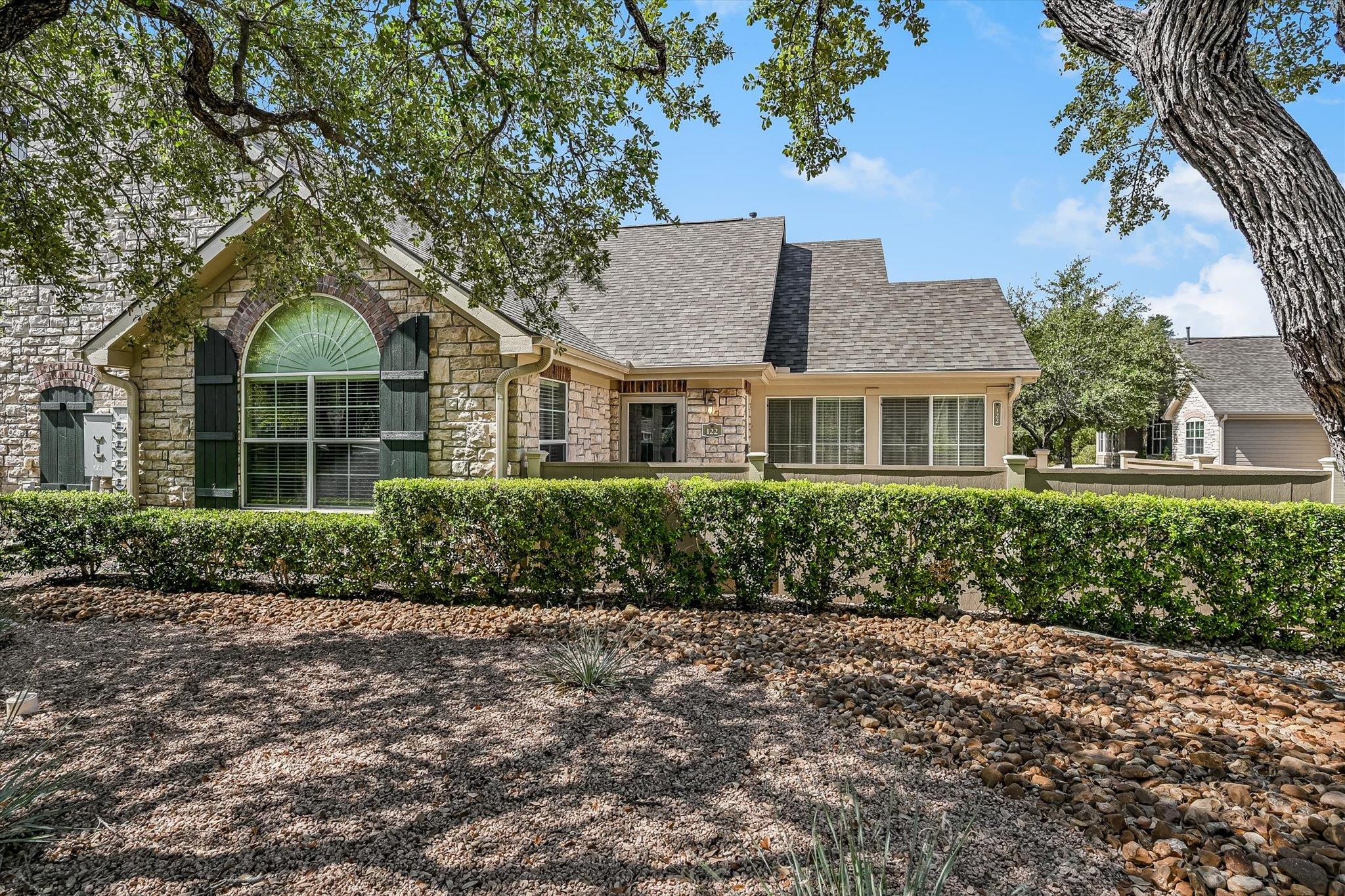 Stone-front home with an arched window, dark shutters, clipped hedges, and rock landscaping.