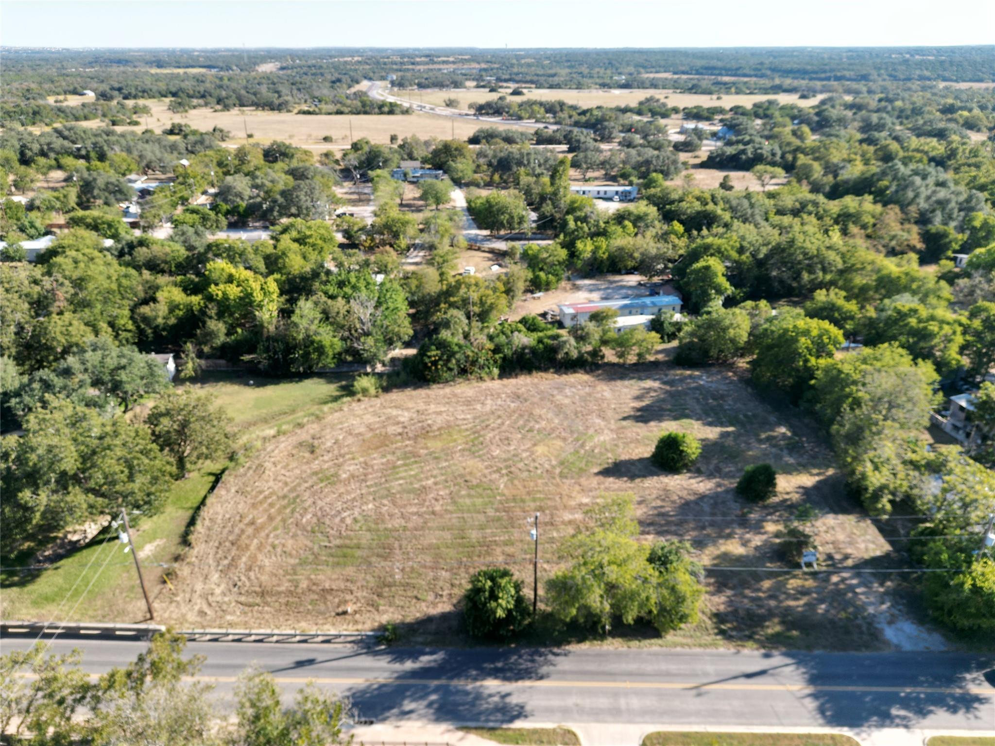 Aerial view of a cleared lot with road frontage, trees, and scattered buildings nearby.