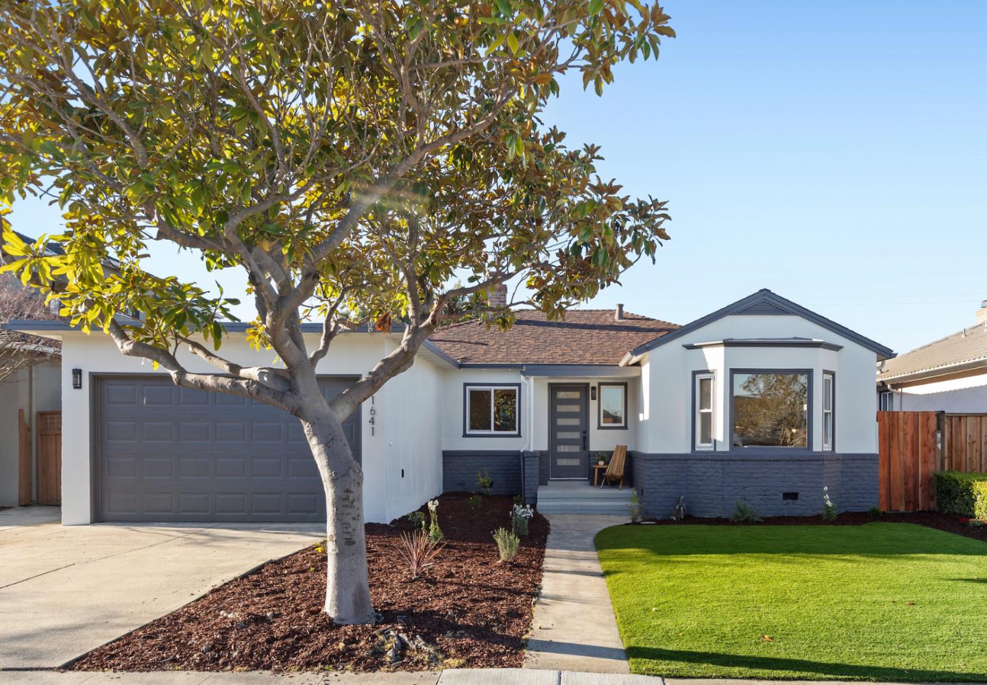 Modern single-story home with a gray garage, projecting bay window, leafy tree, and front lawn.