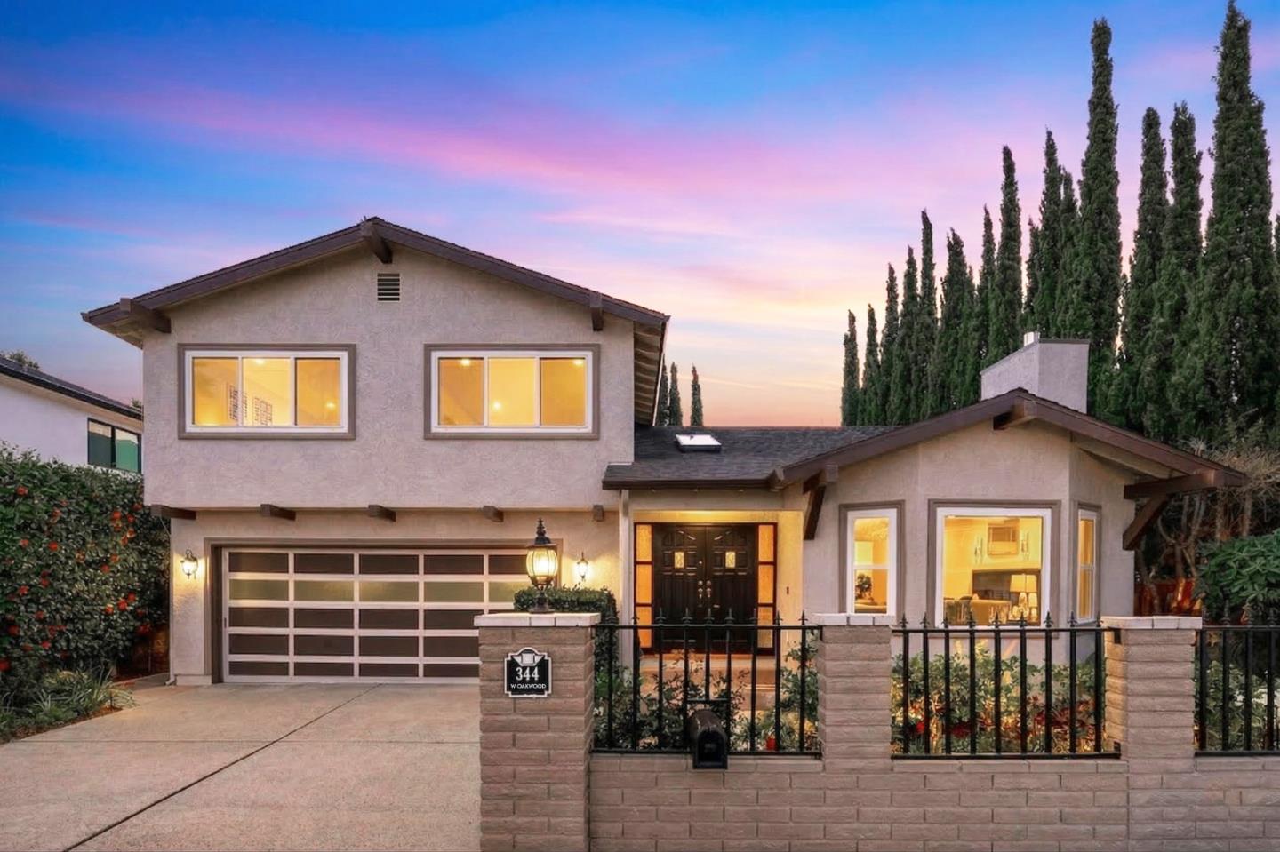 Stucco home with an attached garage, black metal fence, and tall cypress trees at dusk.