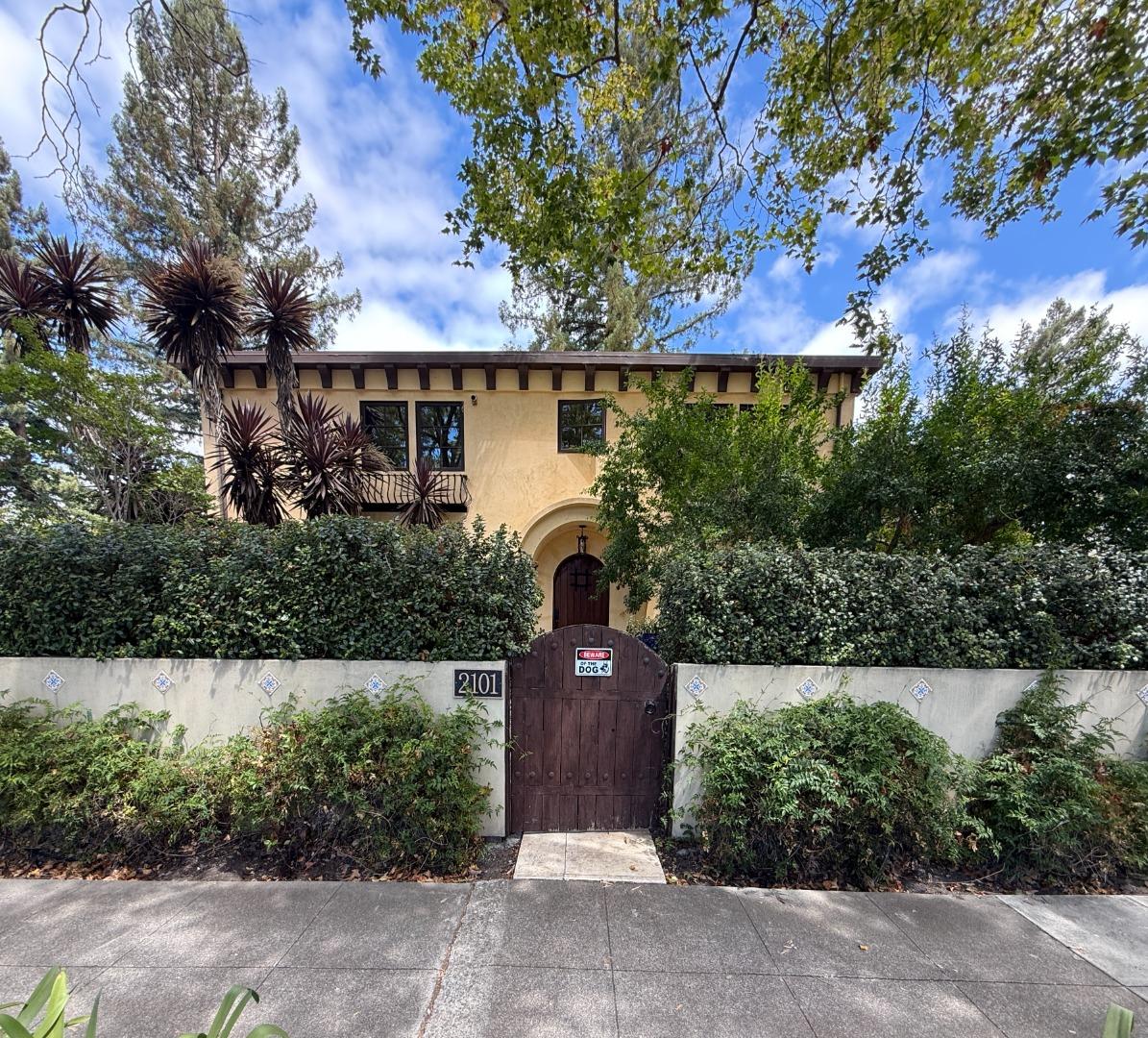 Two-story stucco house behind a wooden arched gate, with address 2101 and dense front hedges.