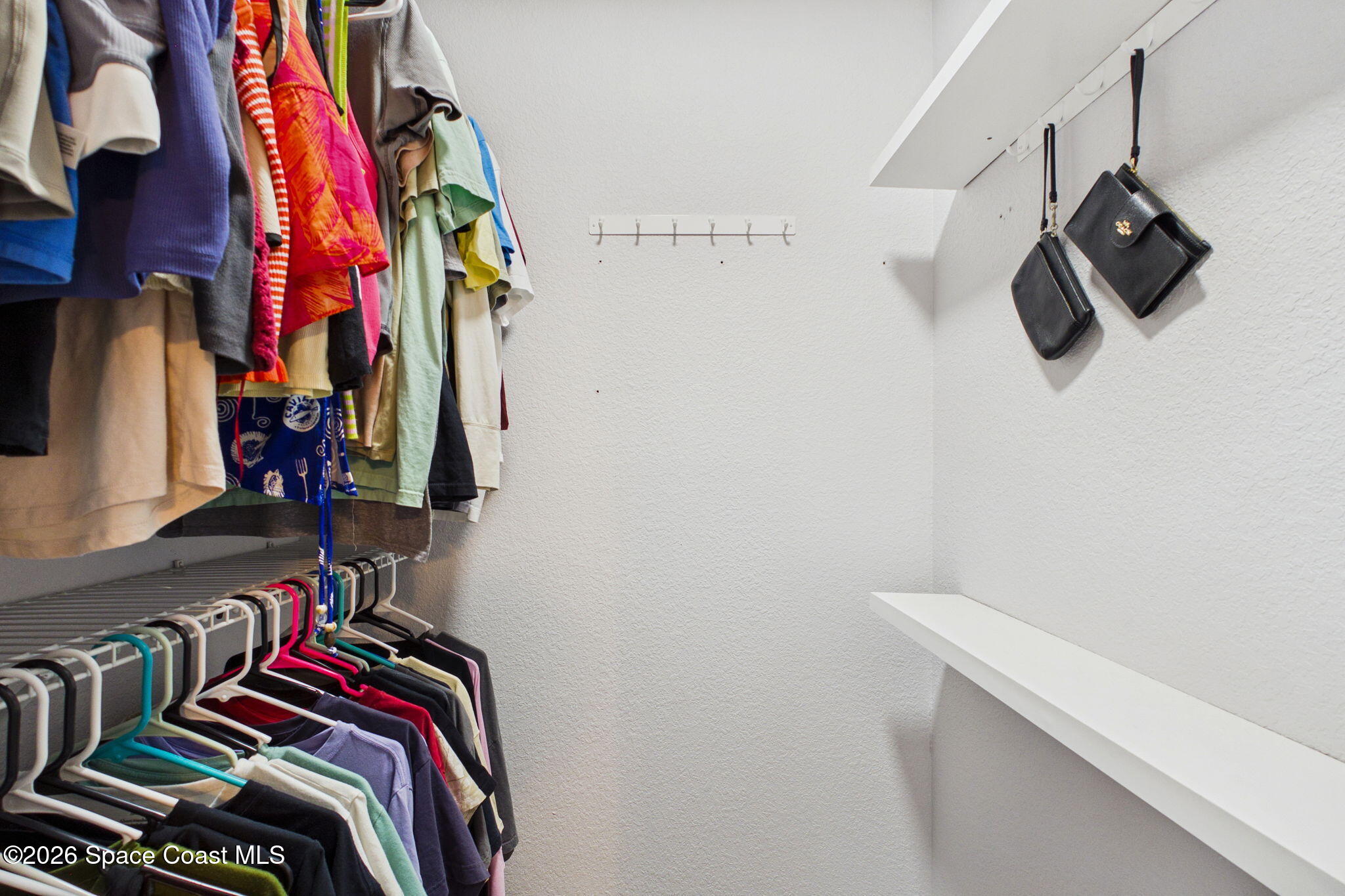 Walk-in closet with clothes on two racks, white shelves, and handbags on wall hooks.