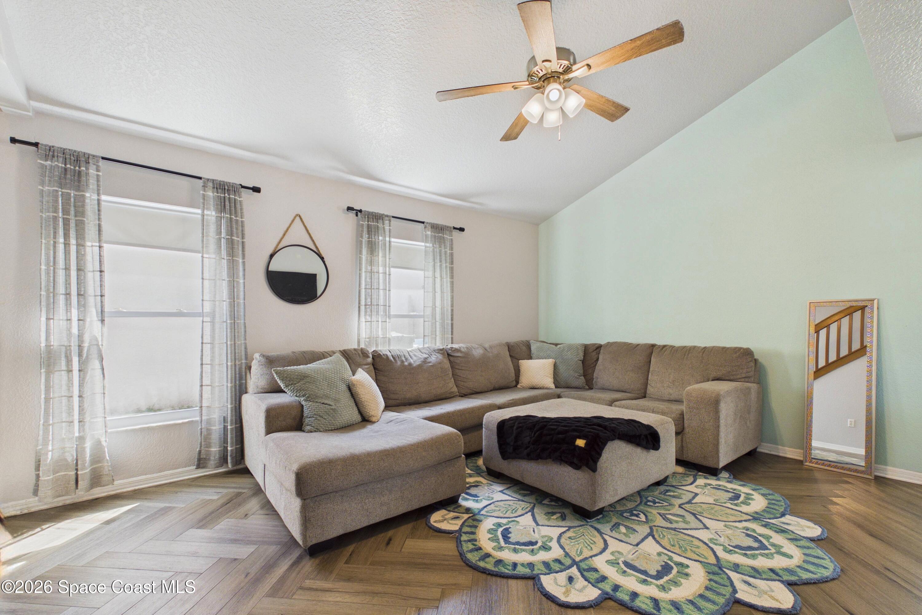 Furnished living room with a gray sectional, patterned rug, pale green accent wall, and ceiling fan.