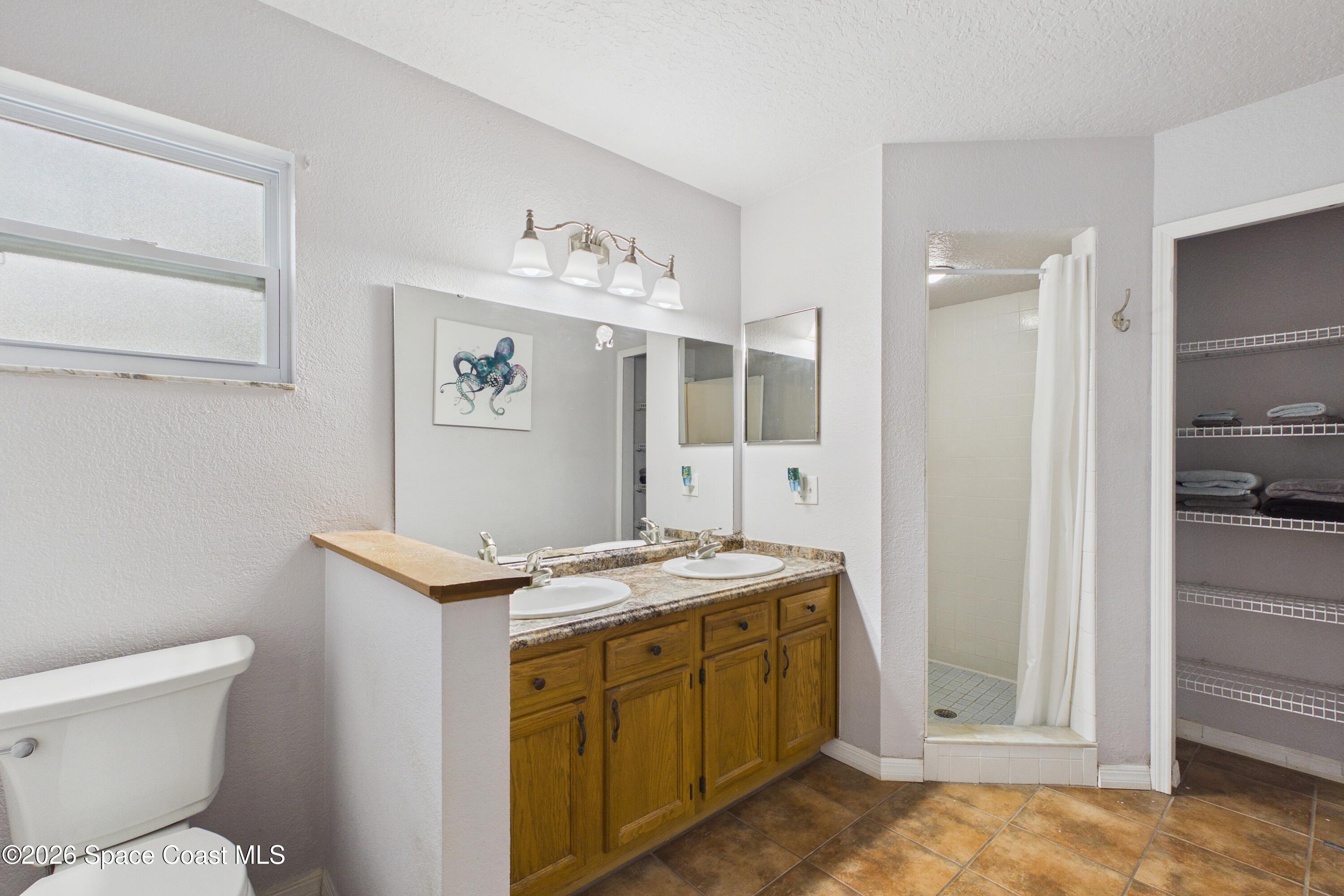 Bathroom with a double-sink wood vanity, tiled shower, open linen shelves, and terracotta-colored tile floor.