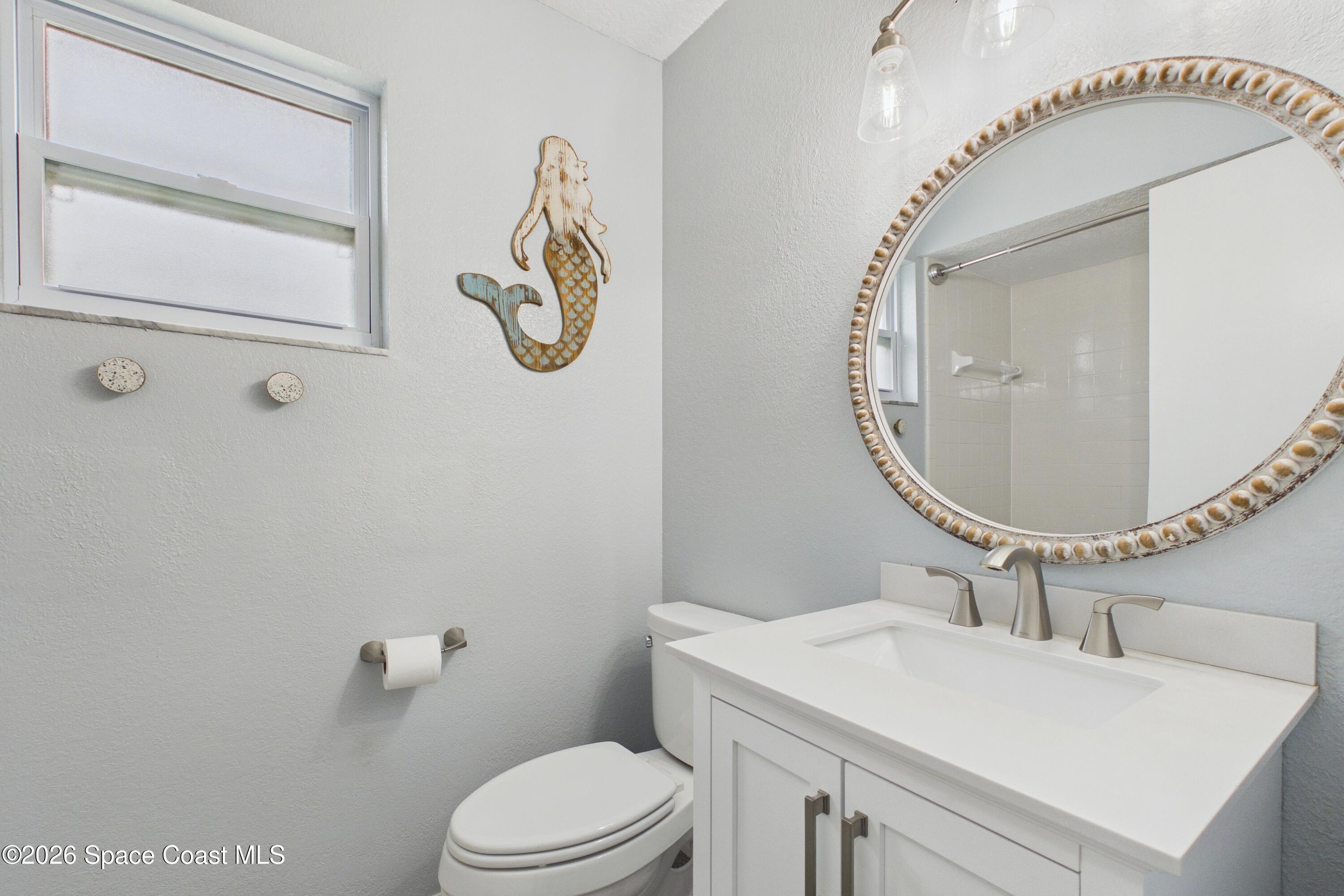 Bathroom with a white vanity, shell-trimmed round mirror, high window, and mermaid wall decoration.