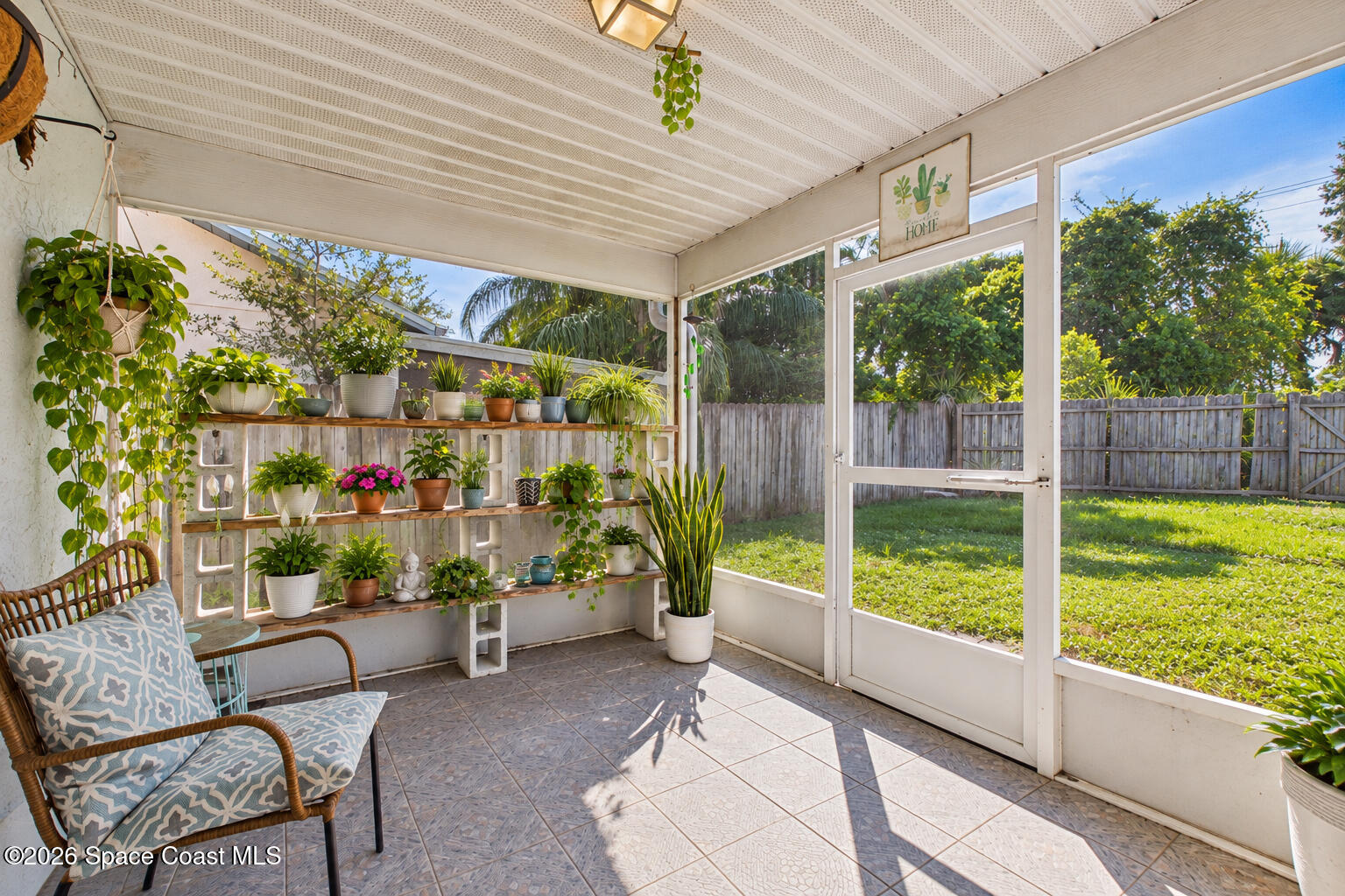 Enclosed sunroom with wicker seating, tiled floor, and shelves of potted plants overlooking a fenced backyard.