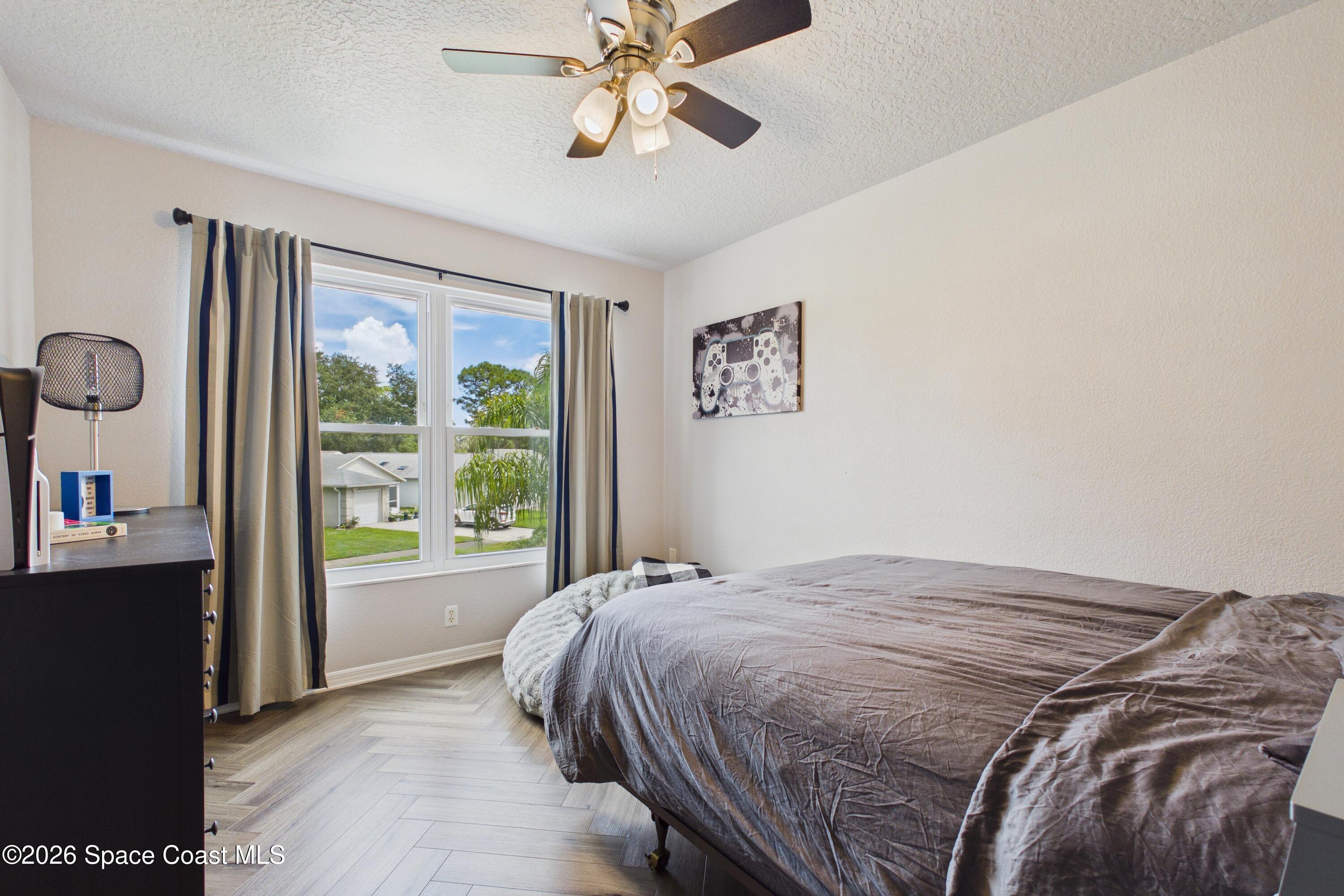 Bedroom with a large gray-covered bed, herringbone flooring, and a window overlooking trees and homes.