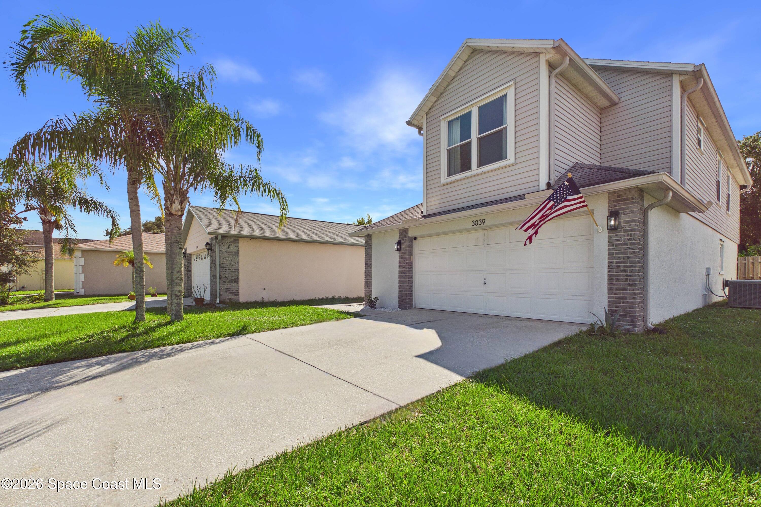 Two-story house with a double garage, American flag, palm trees, and wide driveway.