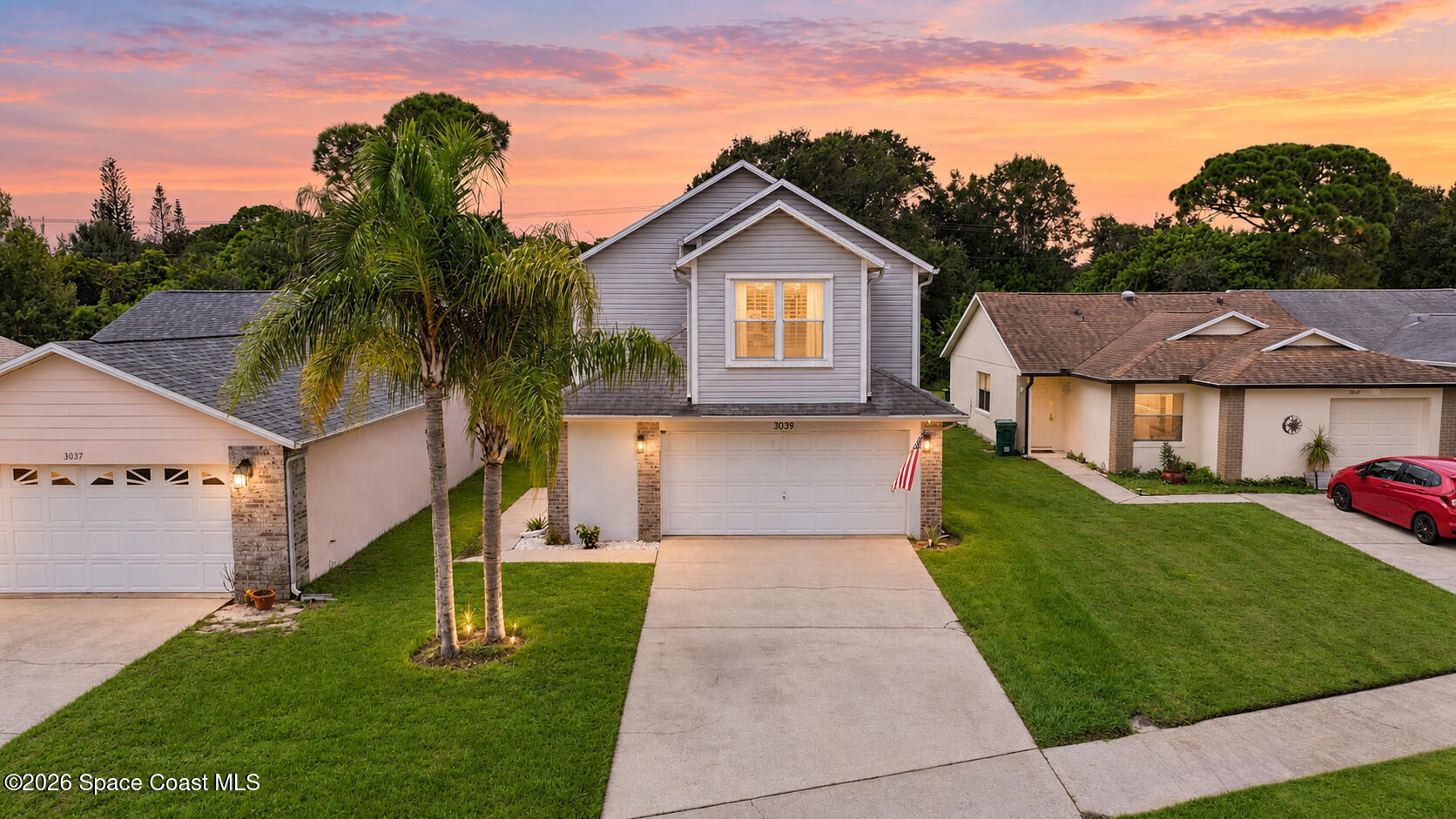Two-story gray house with a double garage, palm trees, and an American flag at sunset.
