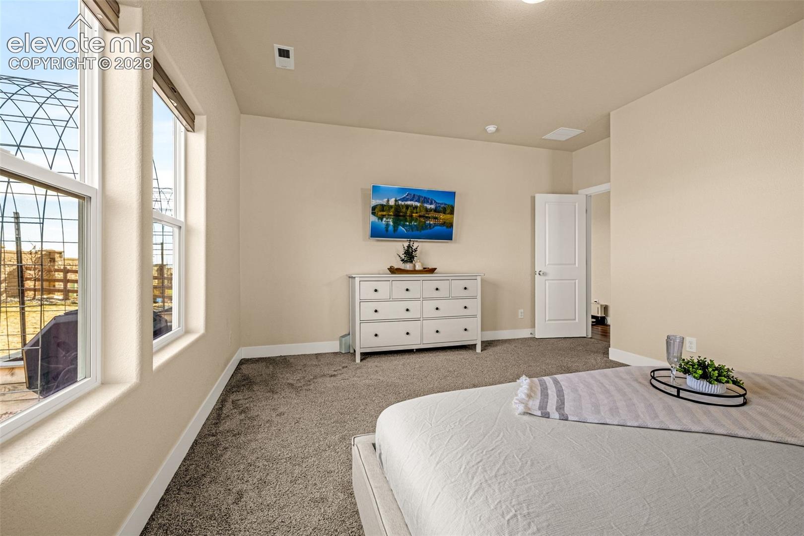 Staged bedroom with tall windows, beige carpet, and a white dresser beside the bed.