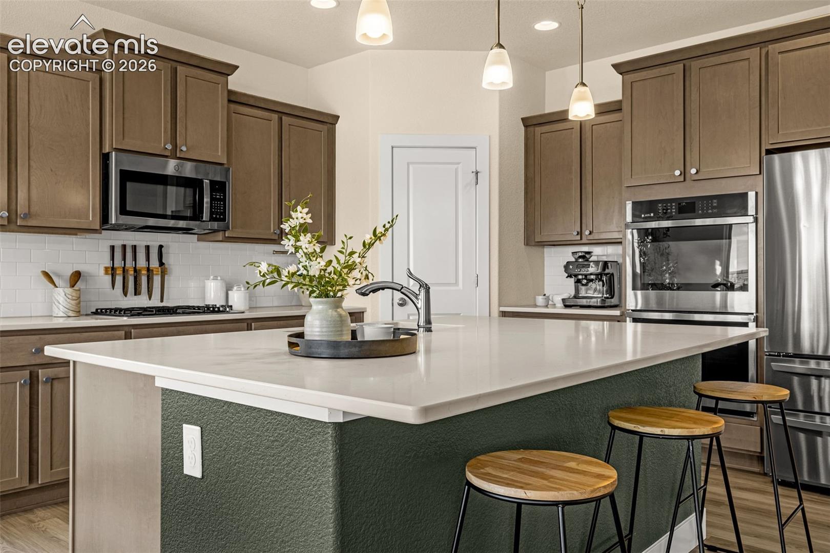 Kitchen with a large white island, dark green base, brown cabinets, and stainless steel appliances.