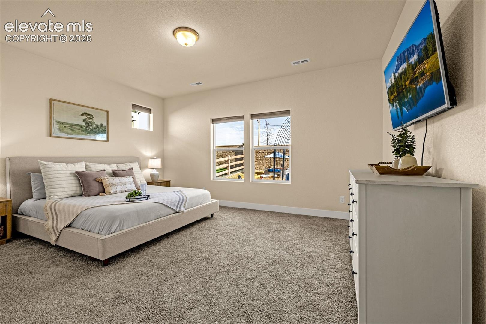 Staged bedroom with a neutral upholstered bed, plush carpet, and wall-mounted television.