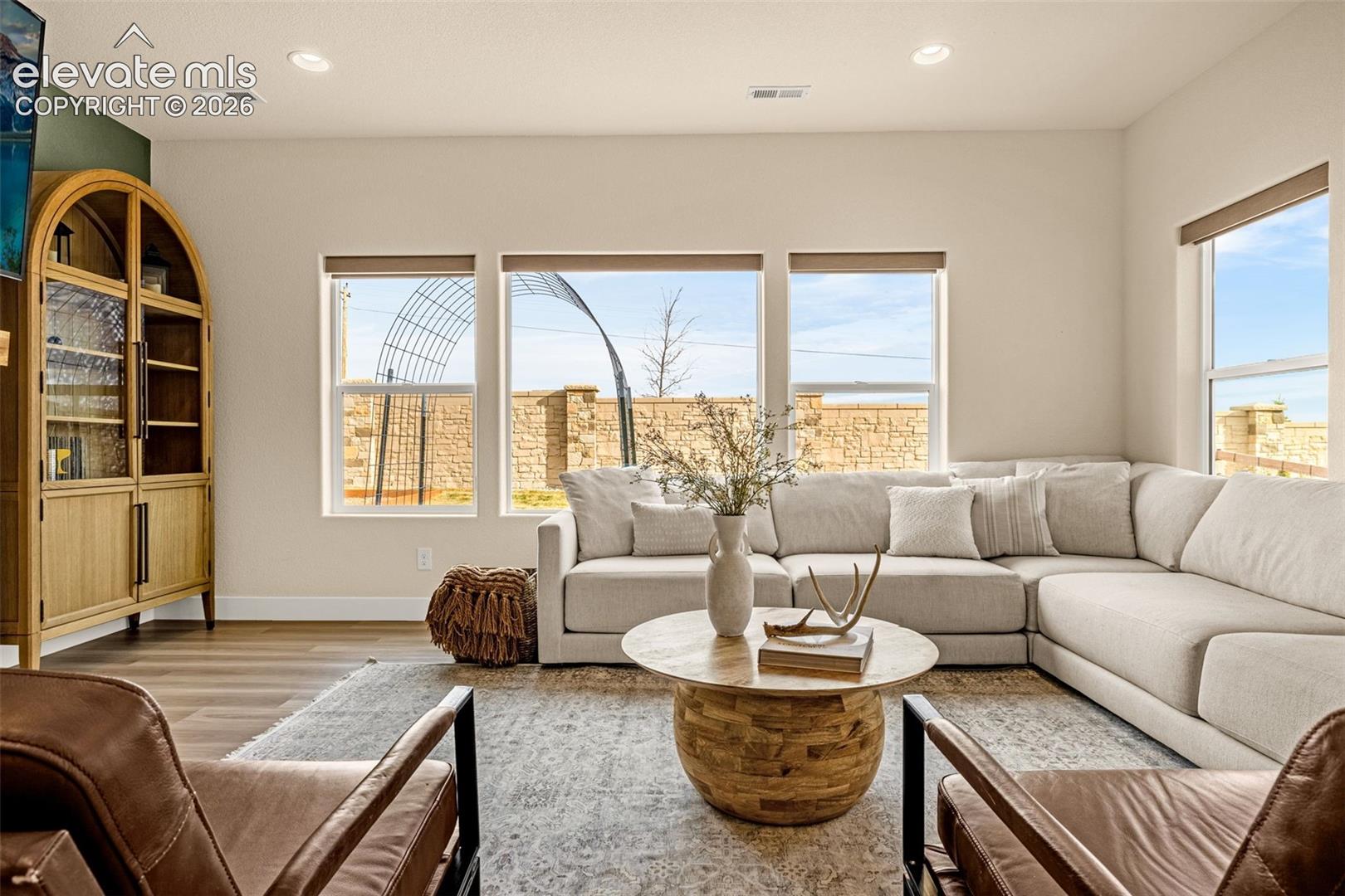 Furnished living room with a cream sectional, leather chairs, and windows overlooking a stone wall.