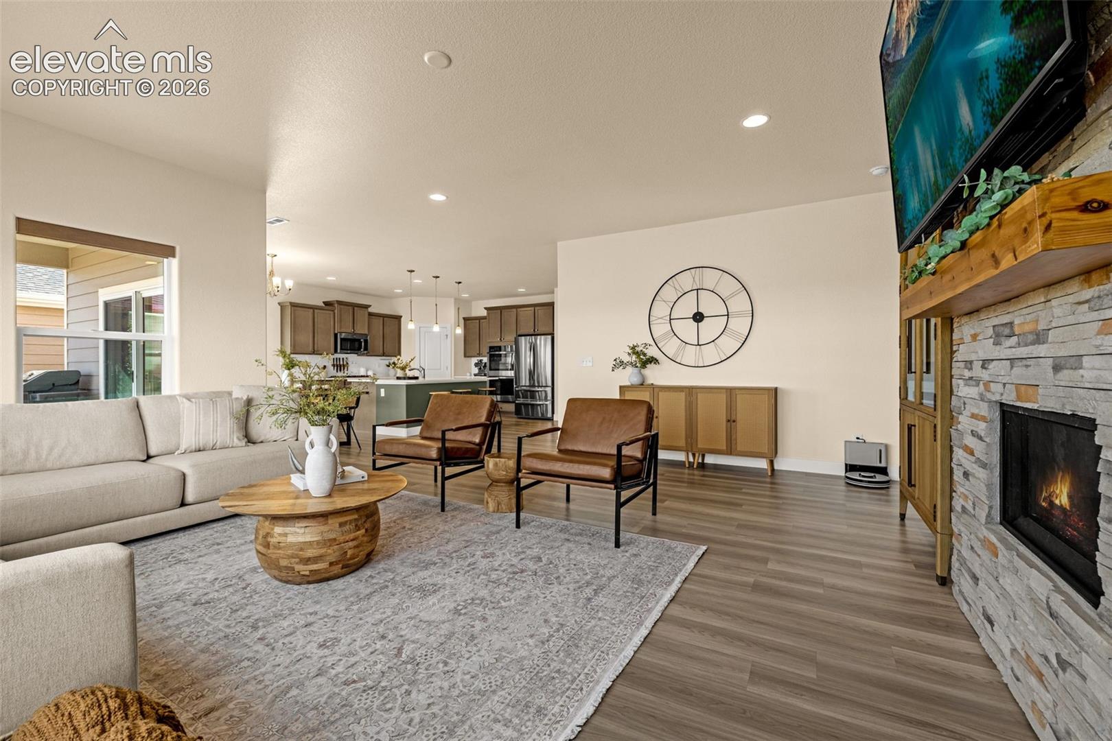 Living room with a stone fireplace, light sectional sofa, and brown leather chairs beside an open kitchen.