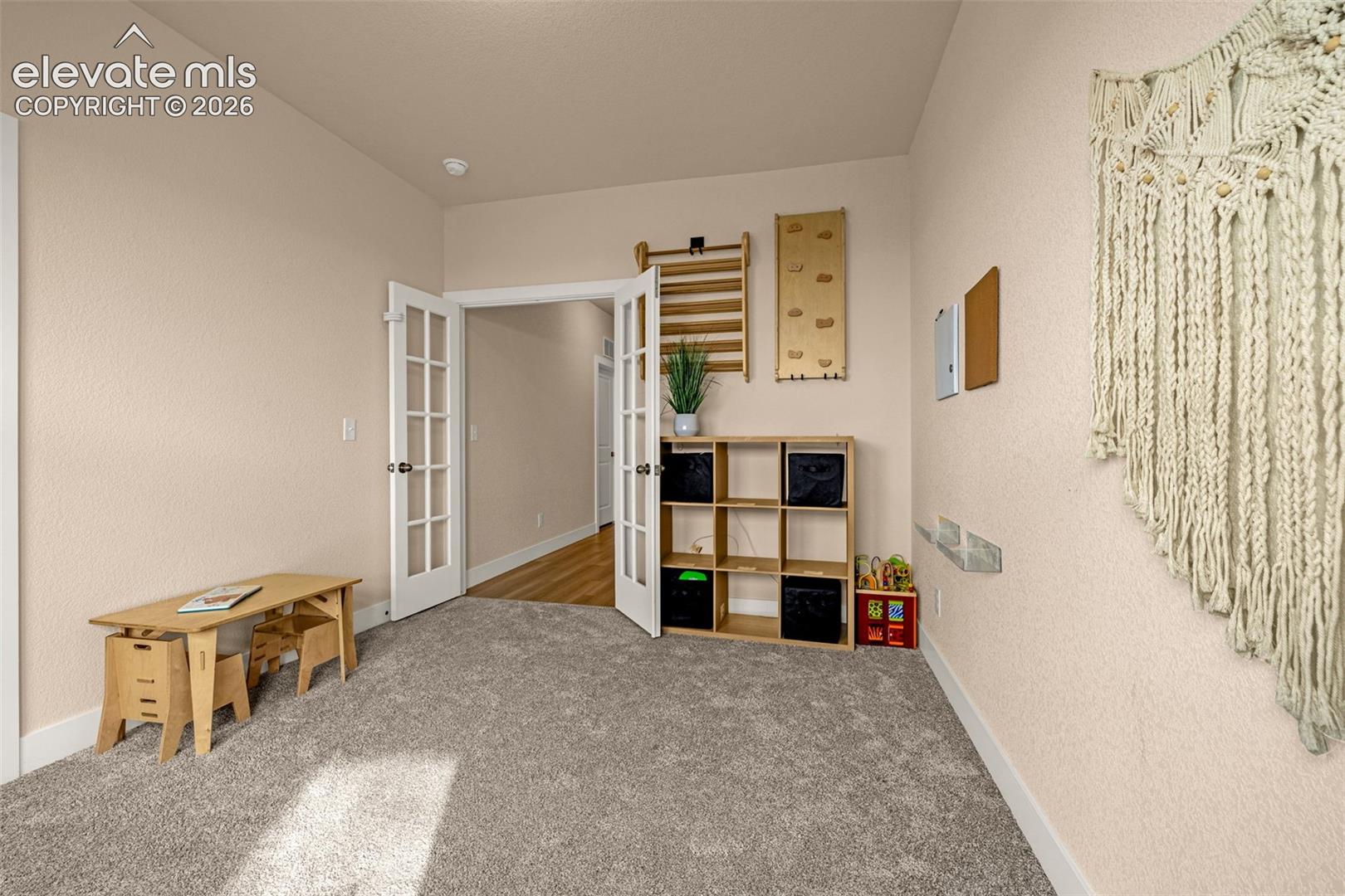 Carpeted playroom with a wooden desk, cubby shelves, toys, and open French doors.