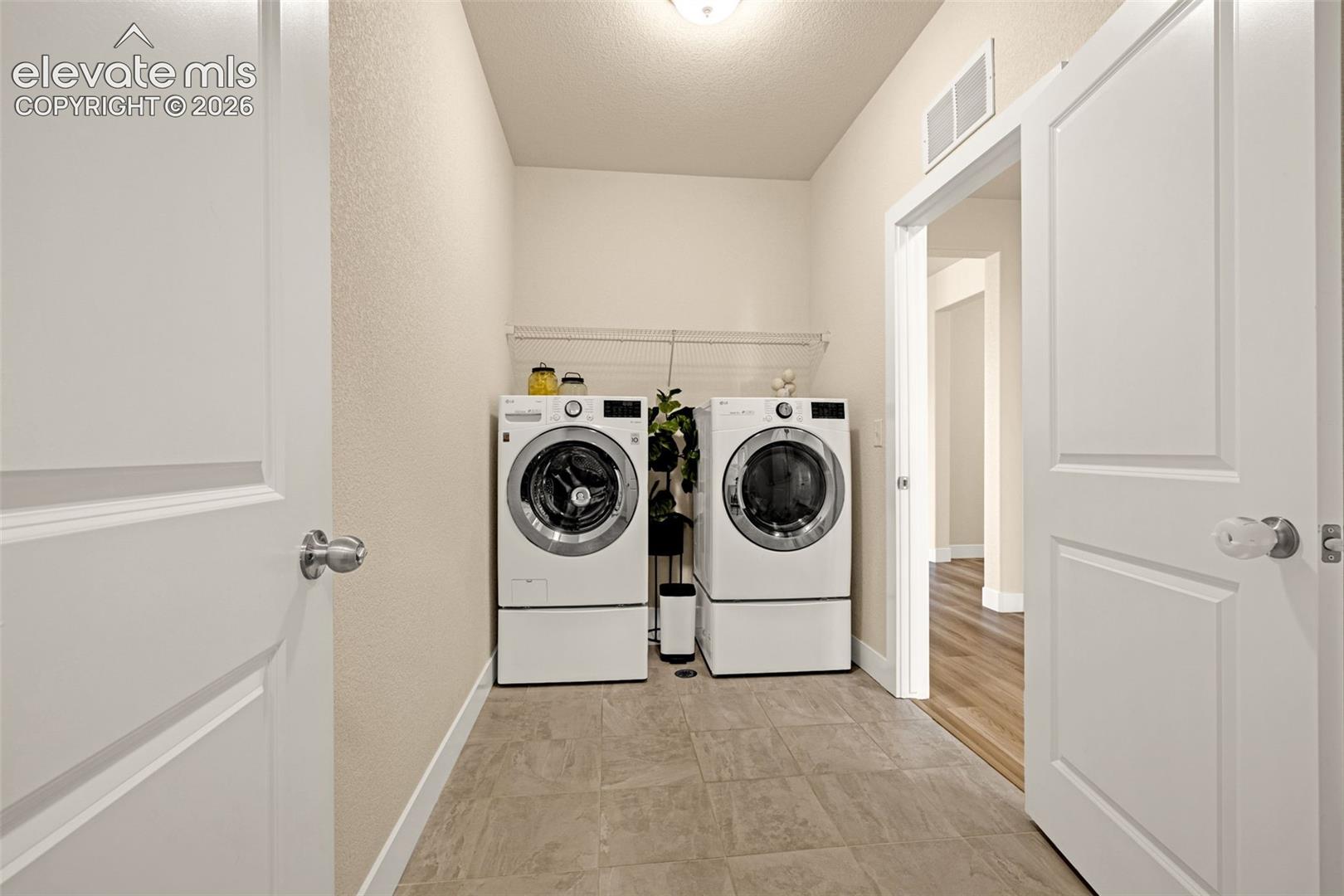 Laundry room with white front-loading washer and dryer, wire shelf, and tiled floor.