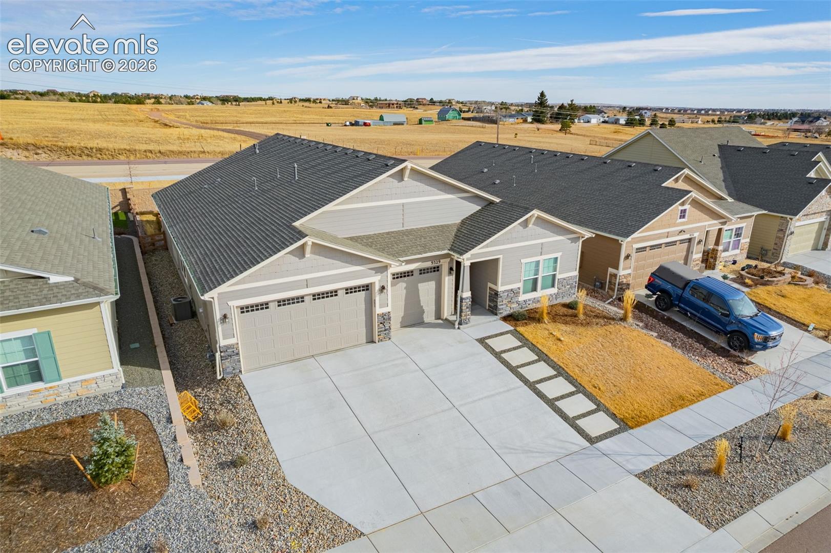 Aerial view of a gray single-story home with stone accents, a wide driveway, and open fields behind it.