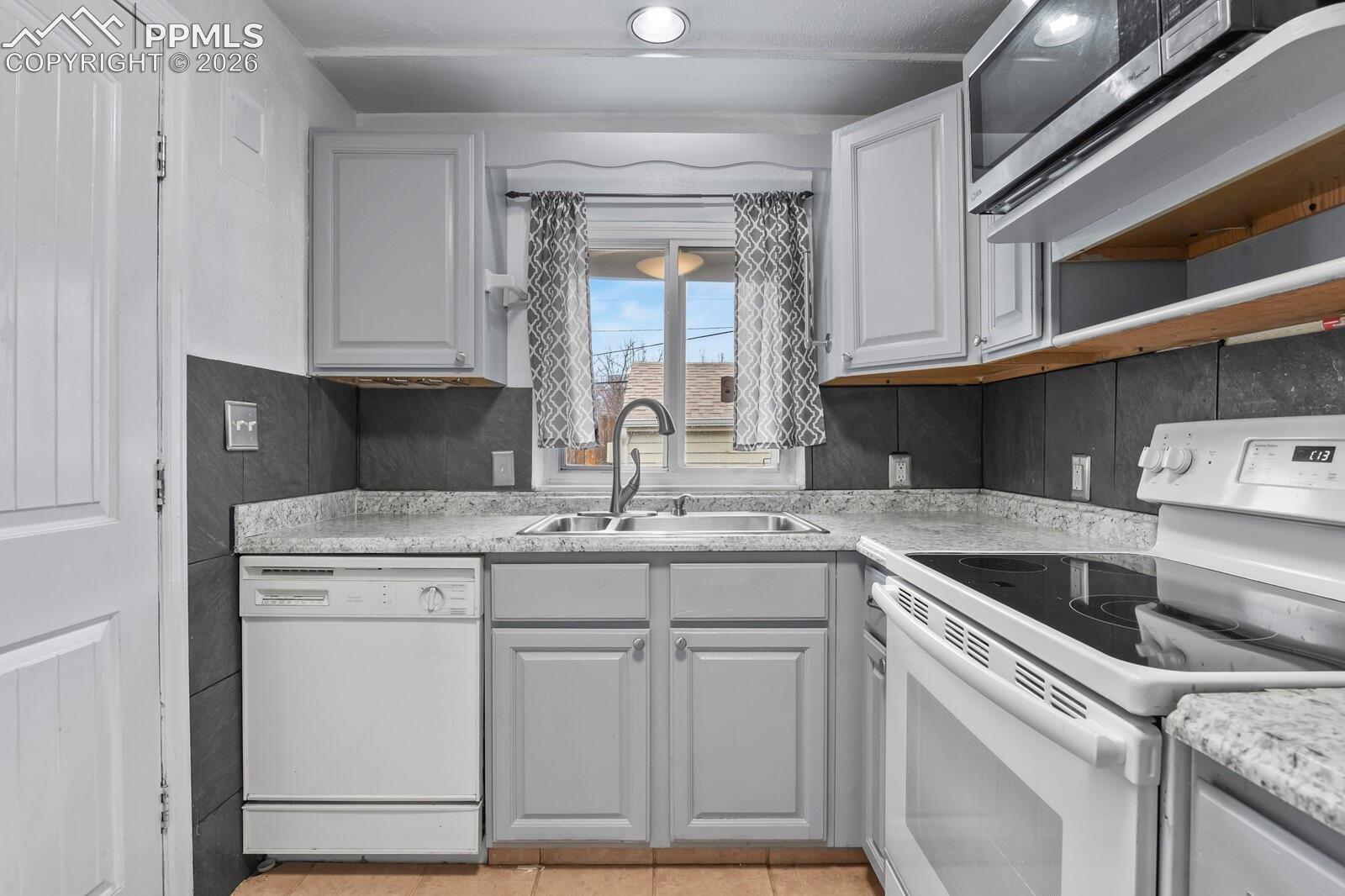 Kitchen with white cabinets, gray tile backsplash, and sink beneath a curtained window.