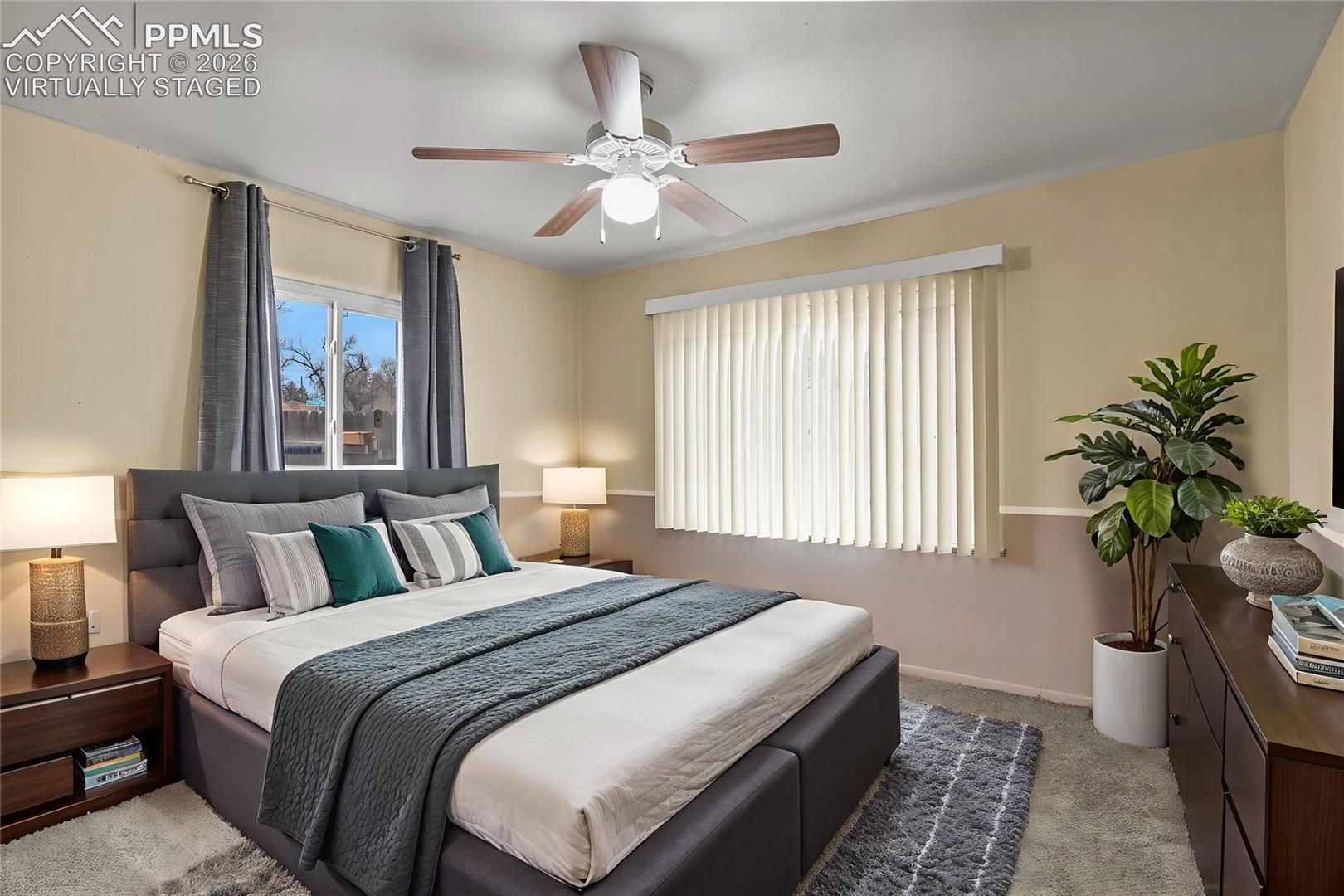 Virtually staged bedroom with a large gray-and-white bed, teal pillows, ceiling fan, and windows with curtains.