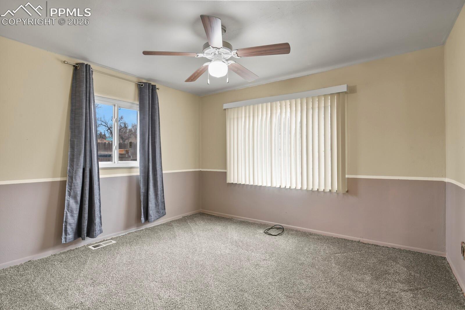 Empty carpeted room with a ceiling fan, gray curtains, and vertical blinds.
