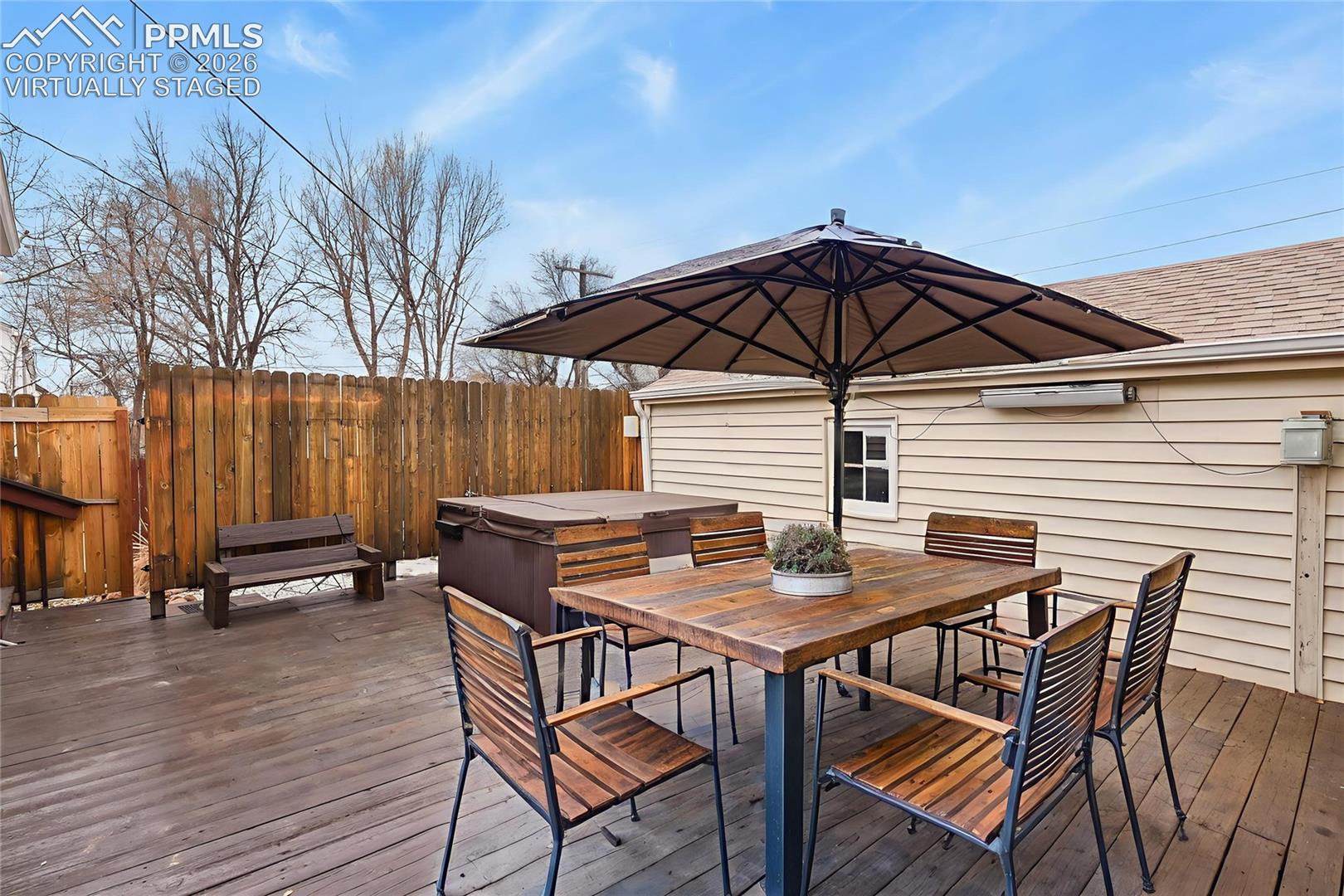 Furnished wooden deck with a hot tub, dining table, large umbrella, and tall privacy fence.