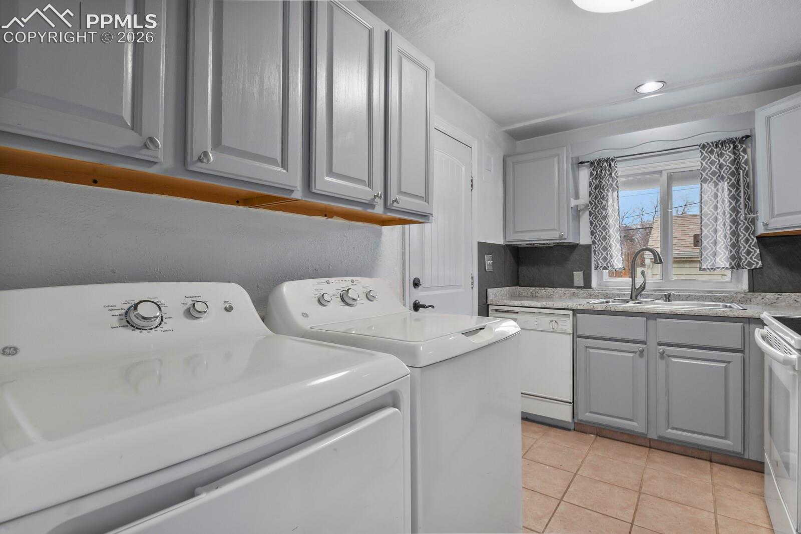 Laundry area with white washer and dryer beside gray kitchen cabinets and a sink under a window.