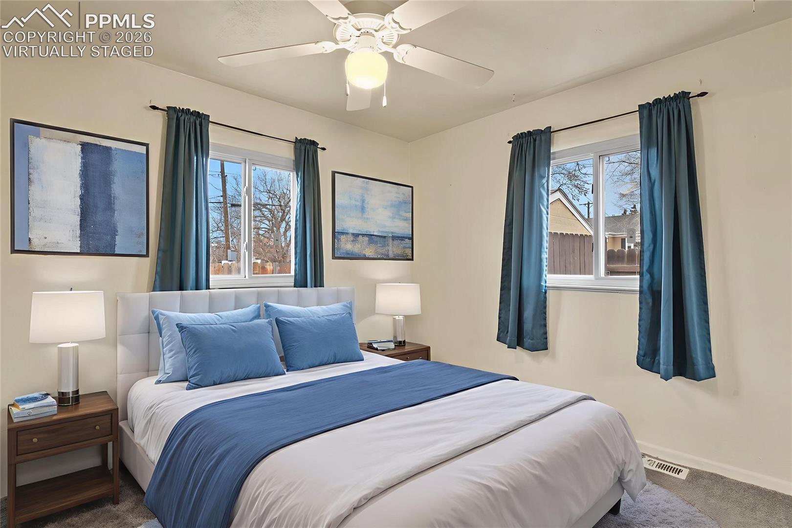 Virtually staged bedroom with a white bed, blue bedding and curtains, and two windows.