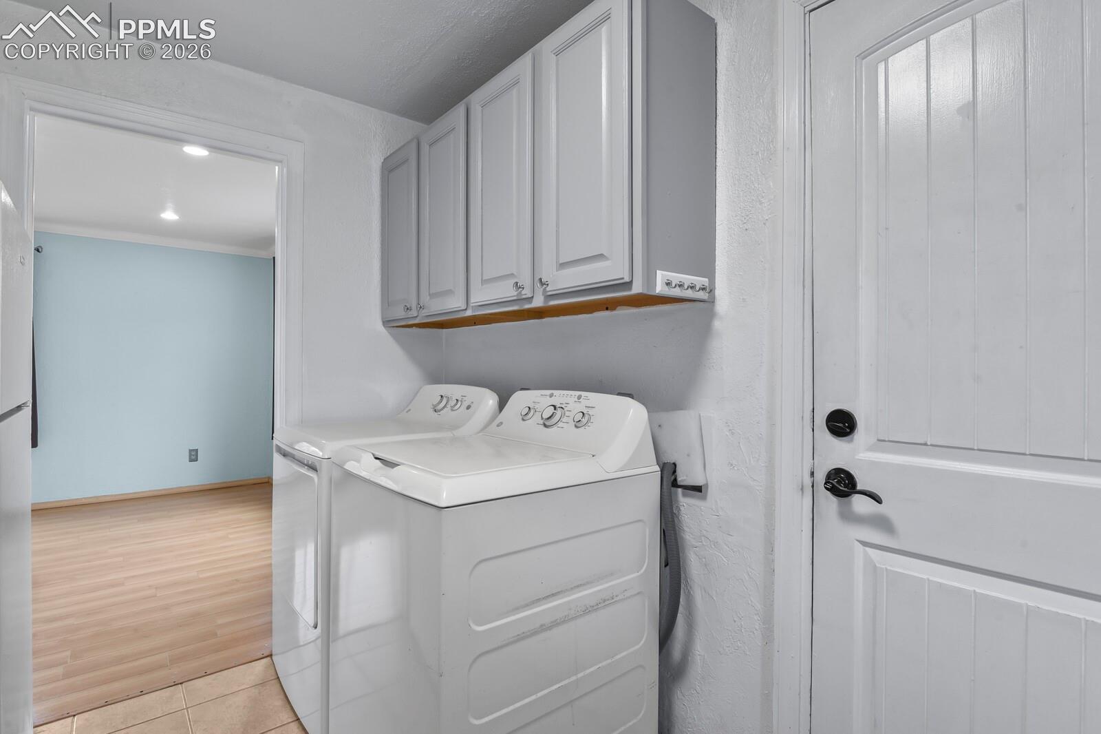 Laundry room with white washer and dryer beneath upper cabinets, opening to a room with light wood-look flooring.