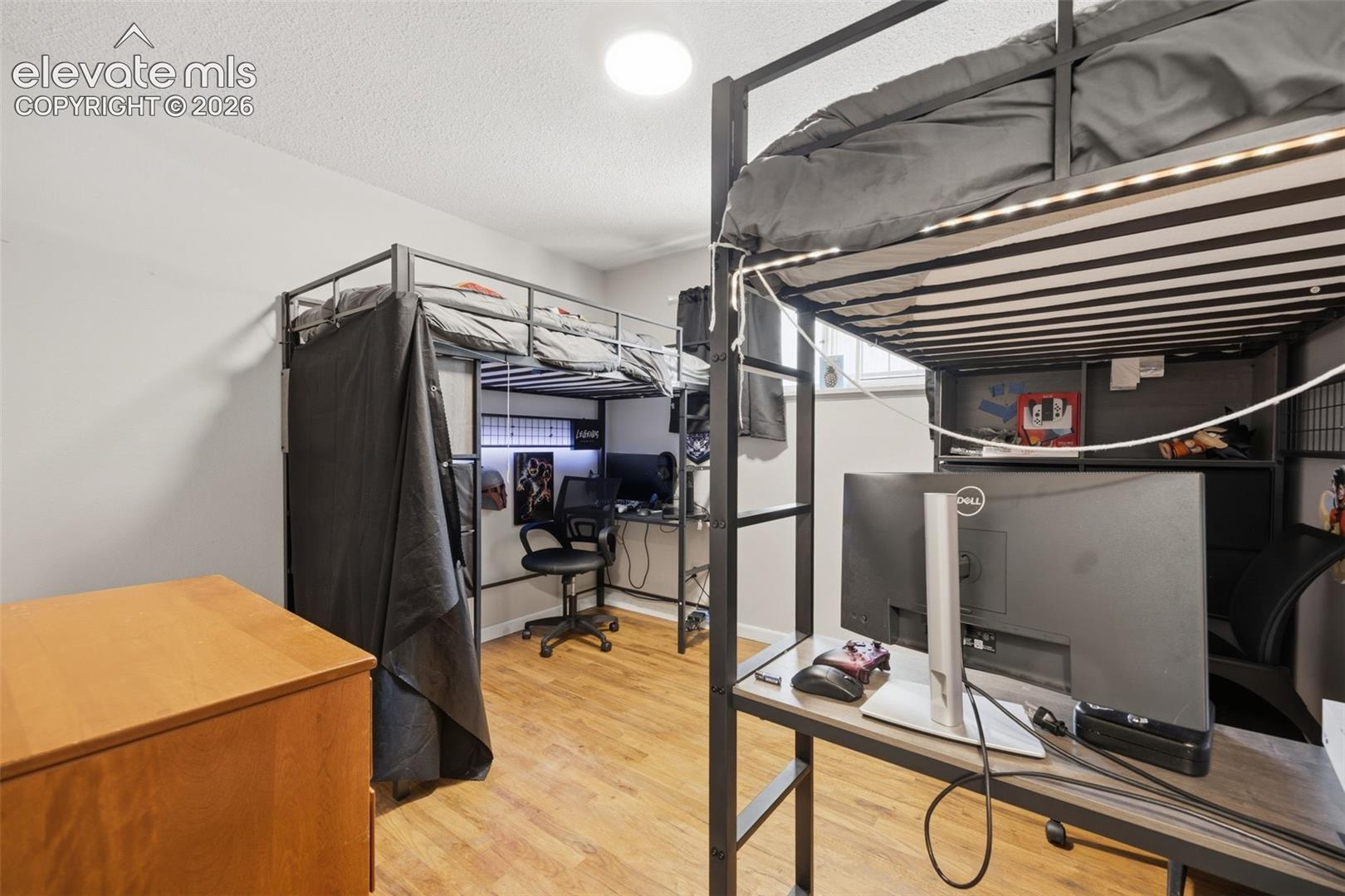 Bedroom with black metal loft beds, computer workstations beneath them, and light wood flooring.