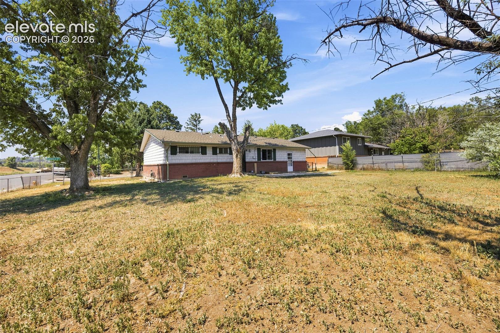 Single-story white-and-brick house behind a dry yard and mature shade trees.
