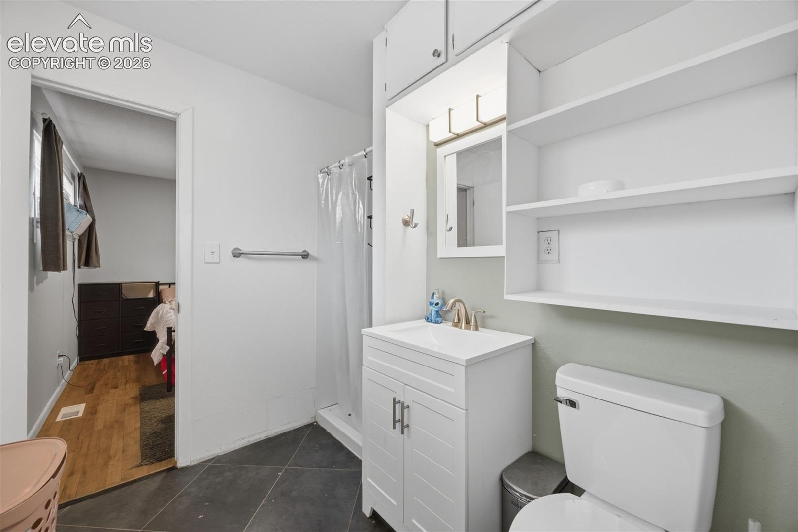 Bathroom with white vanity, toilet, shower curtain, and open shelves beside a bedroom doorway.