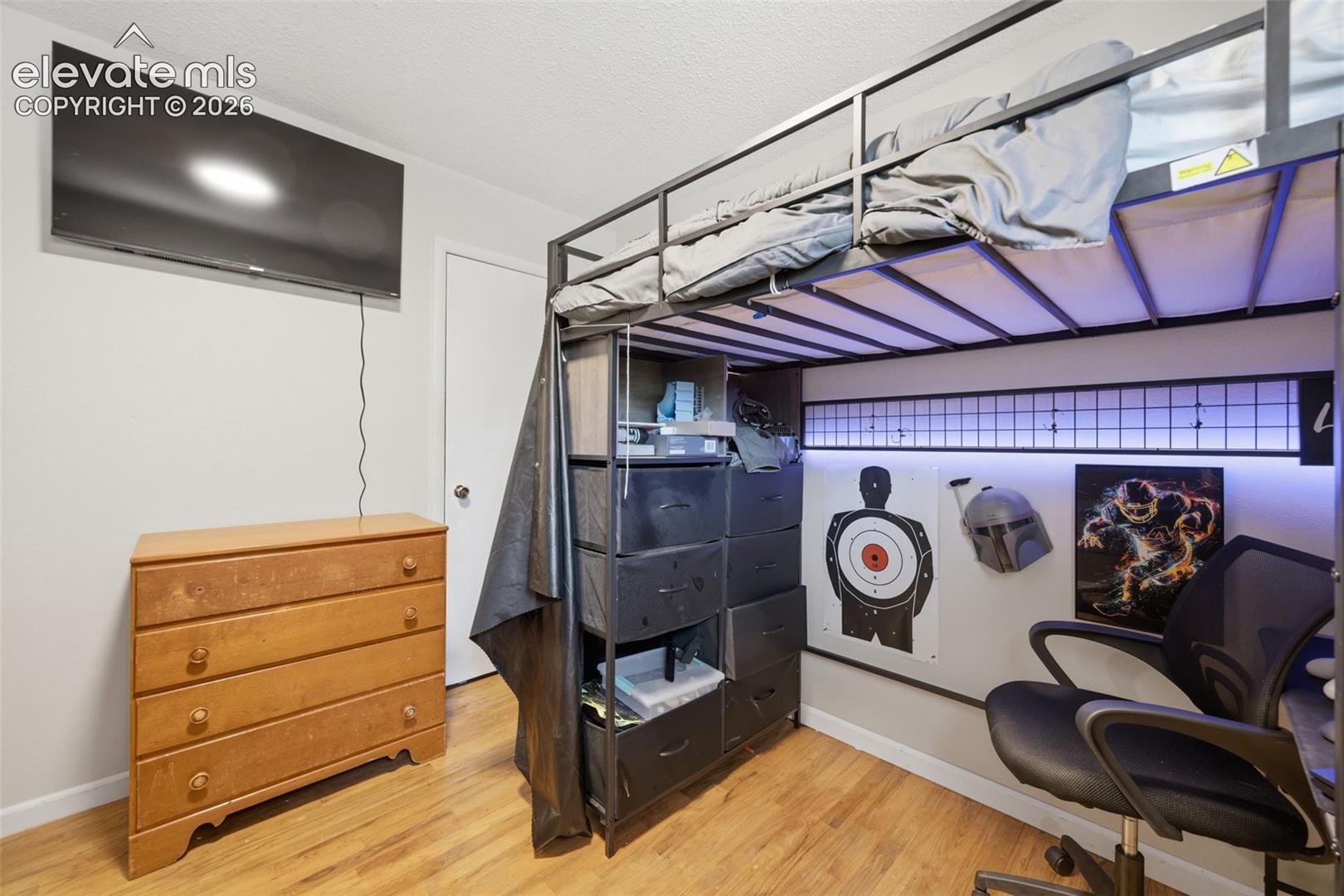 Bedroom with a metal loft bunk bed, wooden dresser, wall-mounted TV, and desk chair.