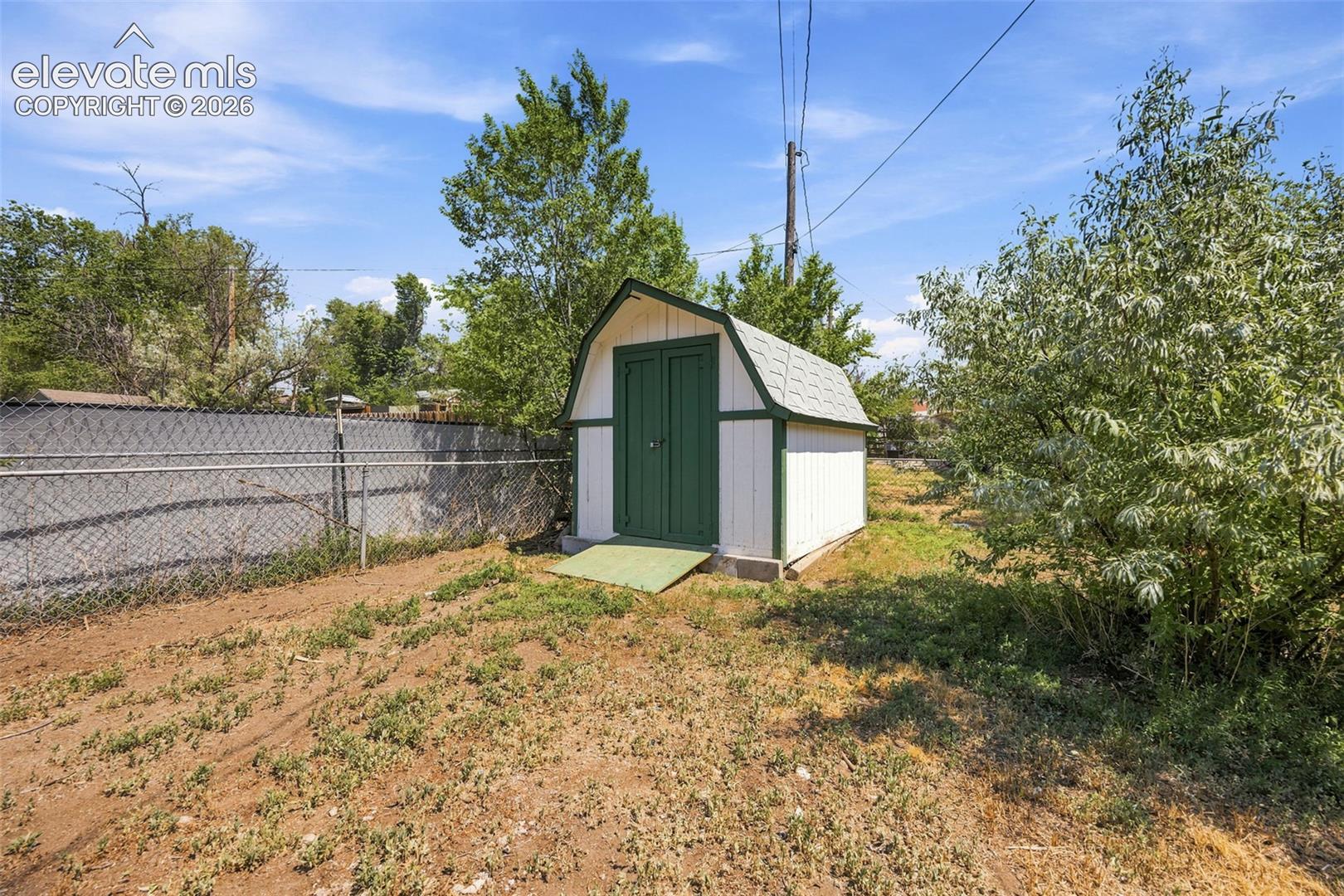 White backyard shed with dark green doors and trim, beside a chain-link fence and leafy trees.