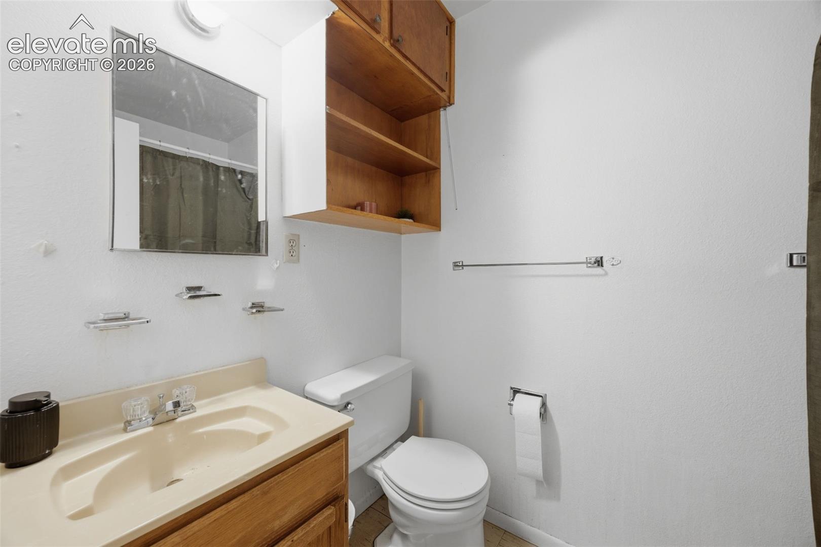 Bathroom with a cream sink, wooden vanity, toilet, and open wall shelving.