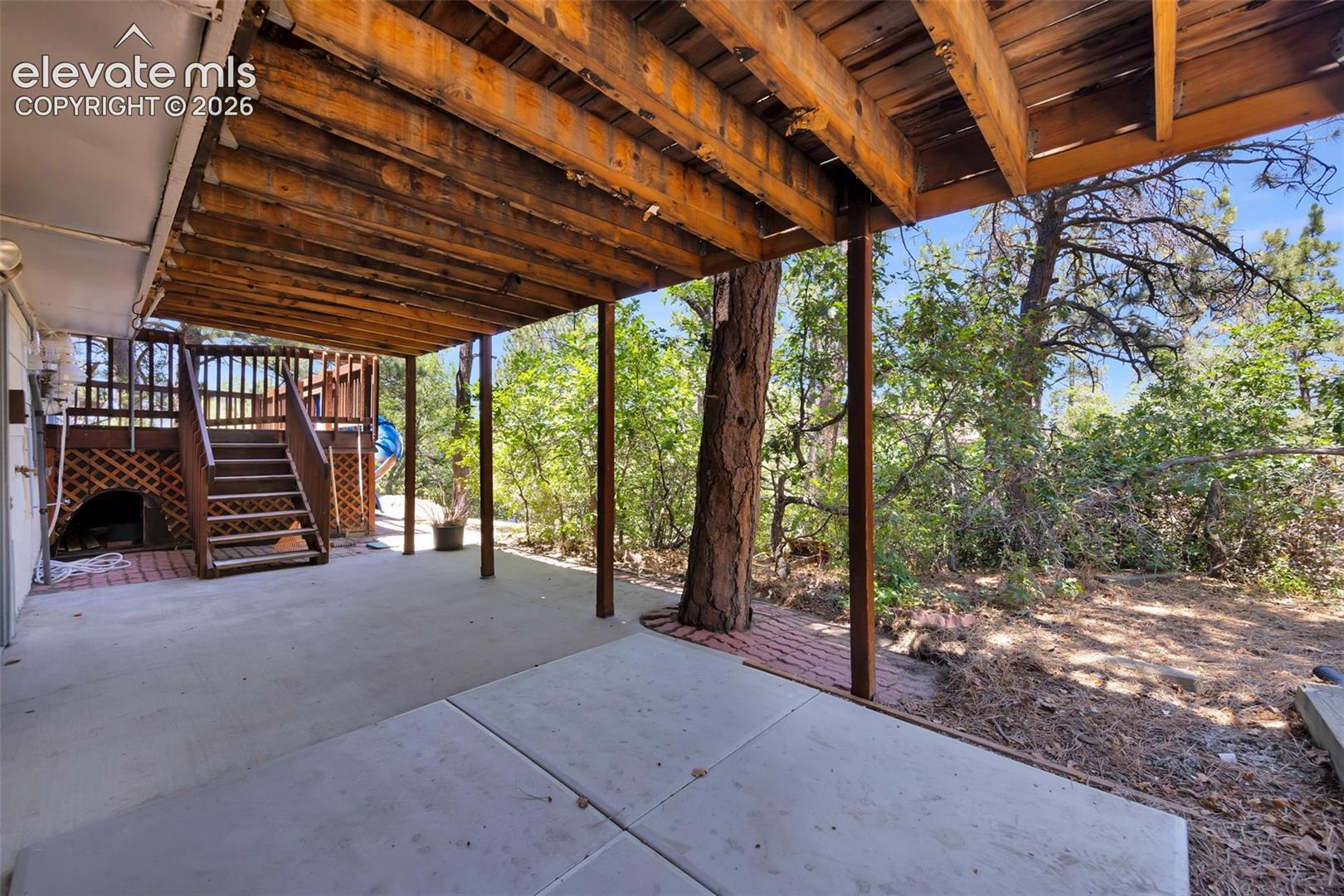 Covered concrete patio with exposed wooden beams, deck stairs, and a mature tree amid wooded landscaping.