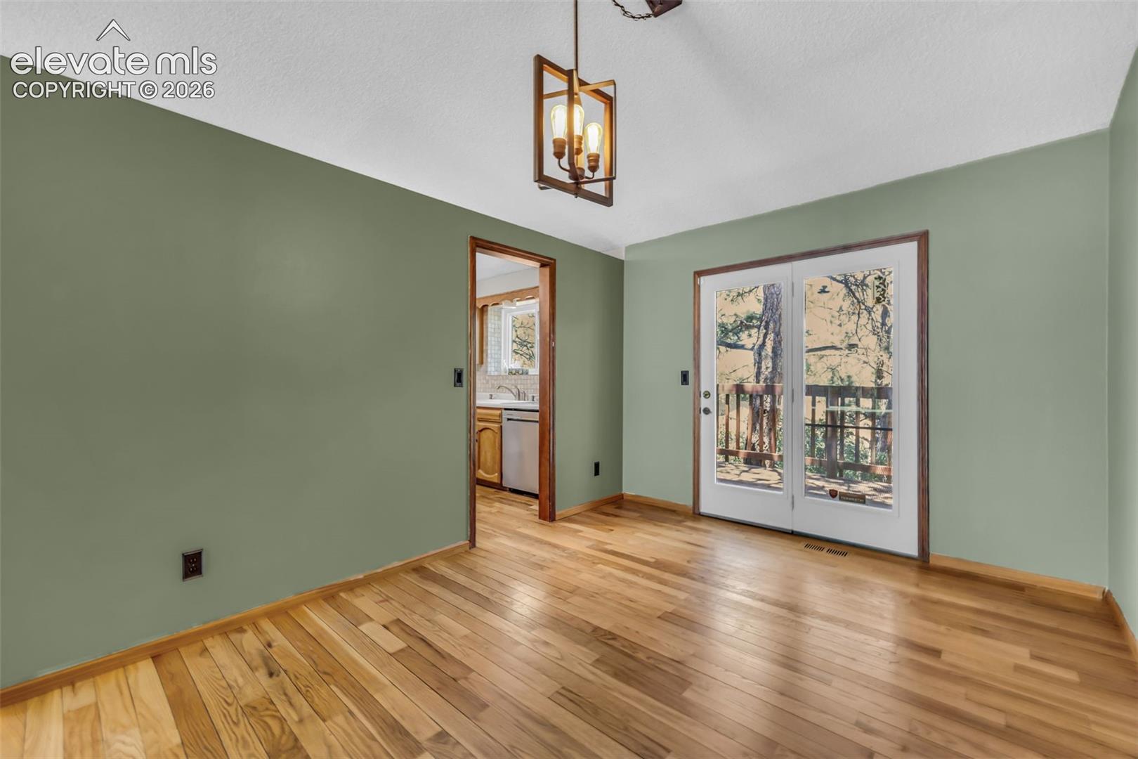 Empty room with hardwood floors, sage-green walls, and glass doors opening to a wooded deck.