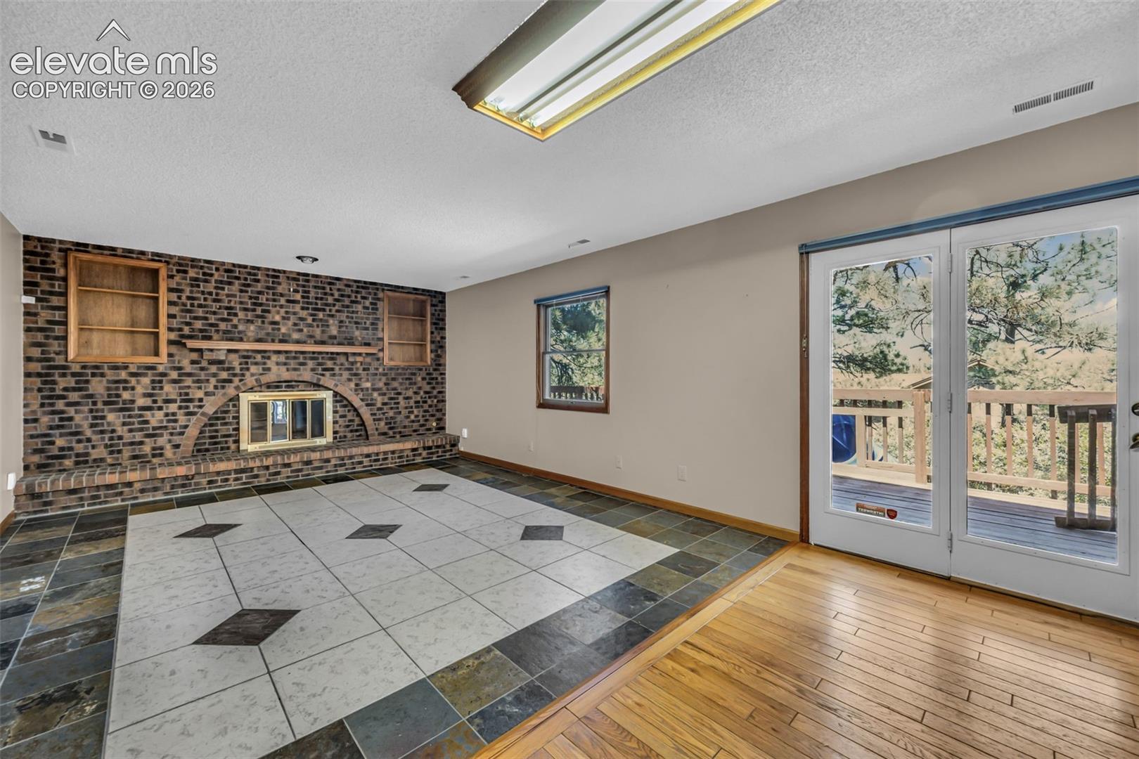 Living room with a brick fireplace, patterned tile floor, and French doors opening to a wooded deck.