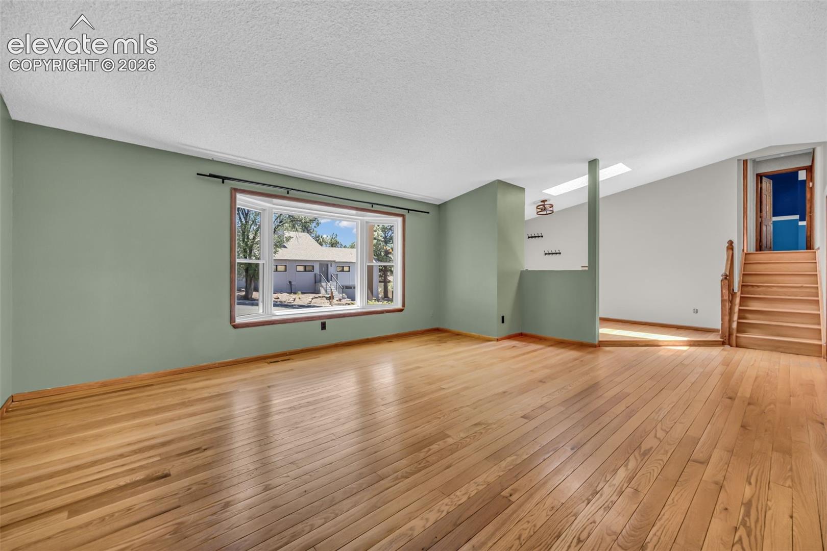 Vacant living room with hardwood floors, sage-green walls, a large window, and a staircase.