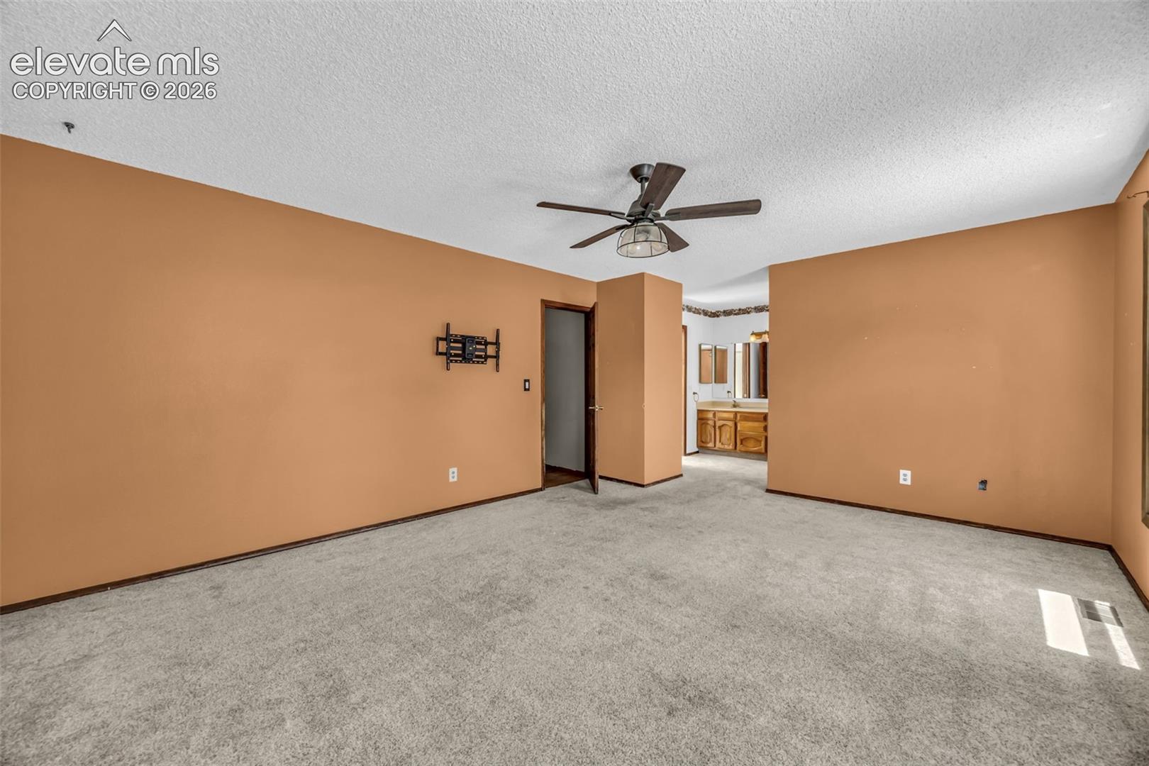 Empty carpeted bedroom with tan walls, a ceiling fan, and an adjoining bathroom vanity.