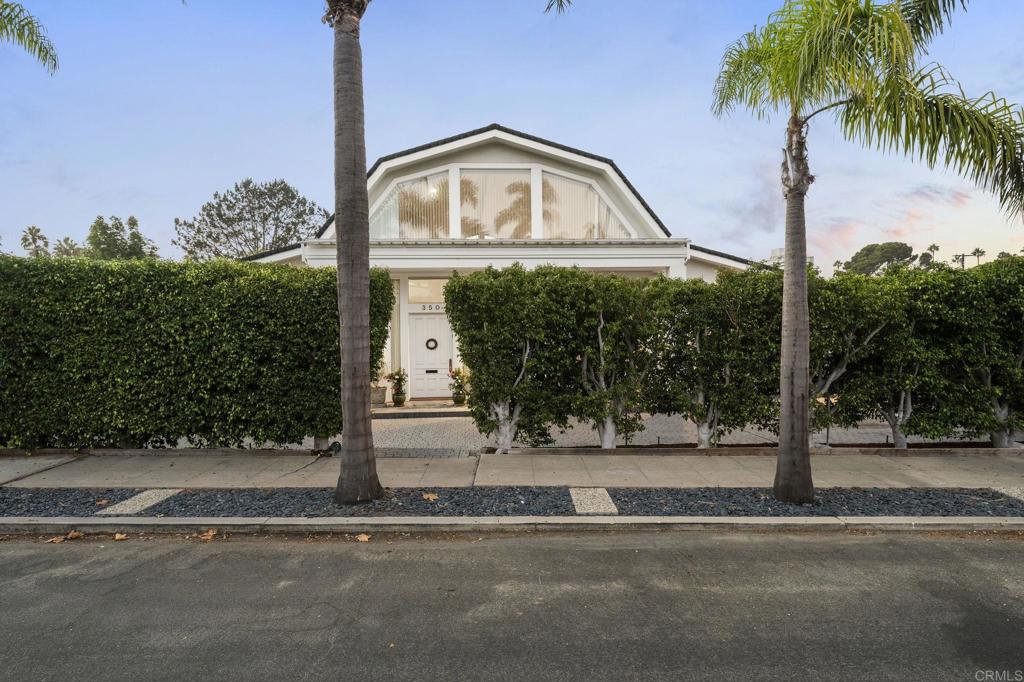 White gabled home with tall hedges and two palm trees along the street.
