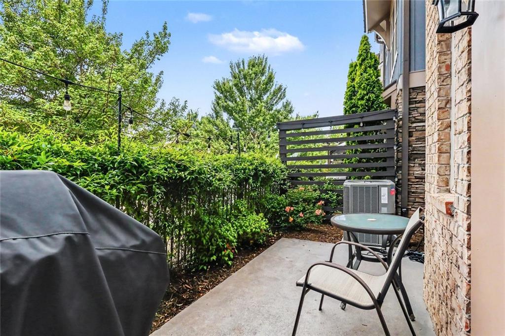 Concrete patio with outdoor seating, a covered grill, string lights, and tall privacy hedges.