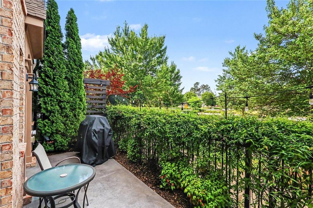 Concrete patio with a glass-top table, covered grill, and leafy hedge beside a brick home.